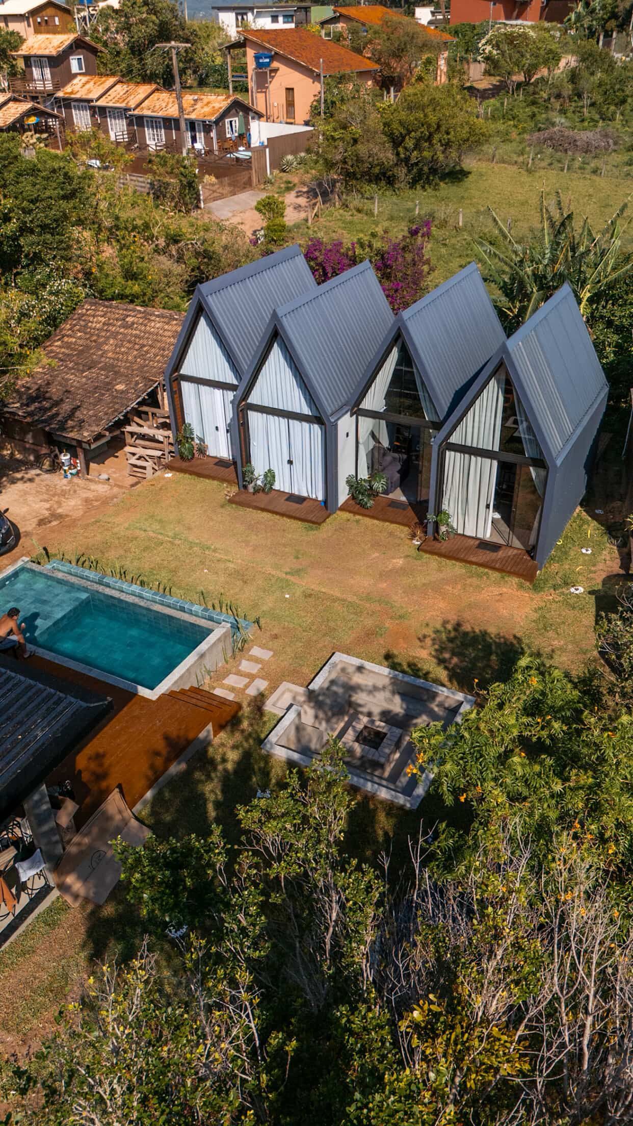 Vista aérea de três cabanas com telhados cinzentos ao lado de uma piscina turquesa, em meio a árvores e terreno rochoso.