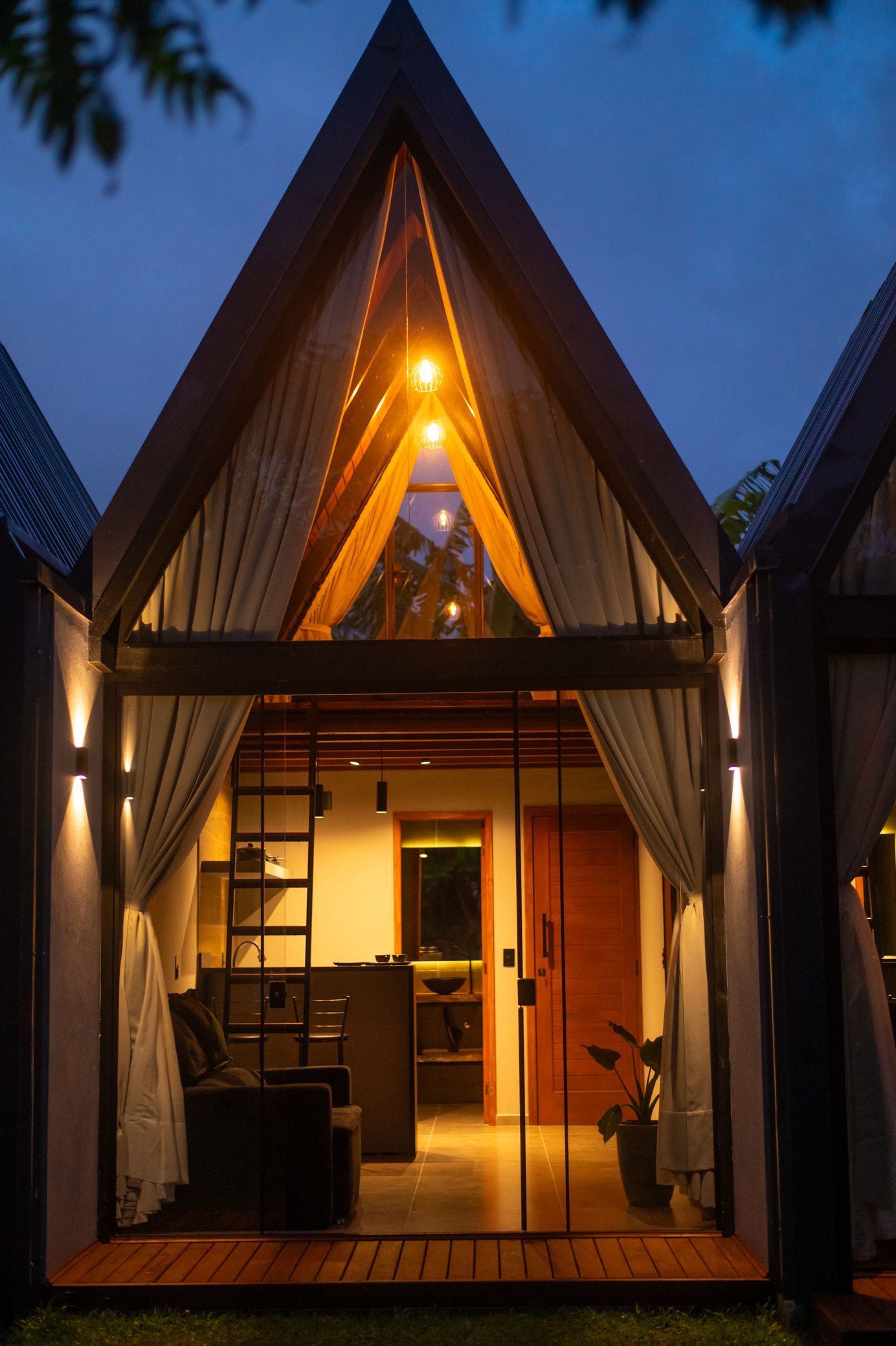 Entrada de uma cabana em forma de A, iluminada de forma aconchegante ao entardecer, com portas abertas e luzes de corda brilhantes no interior.