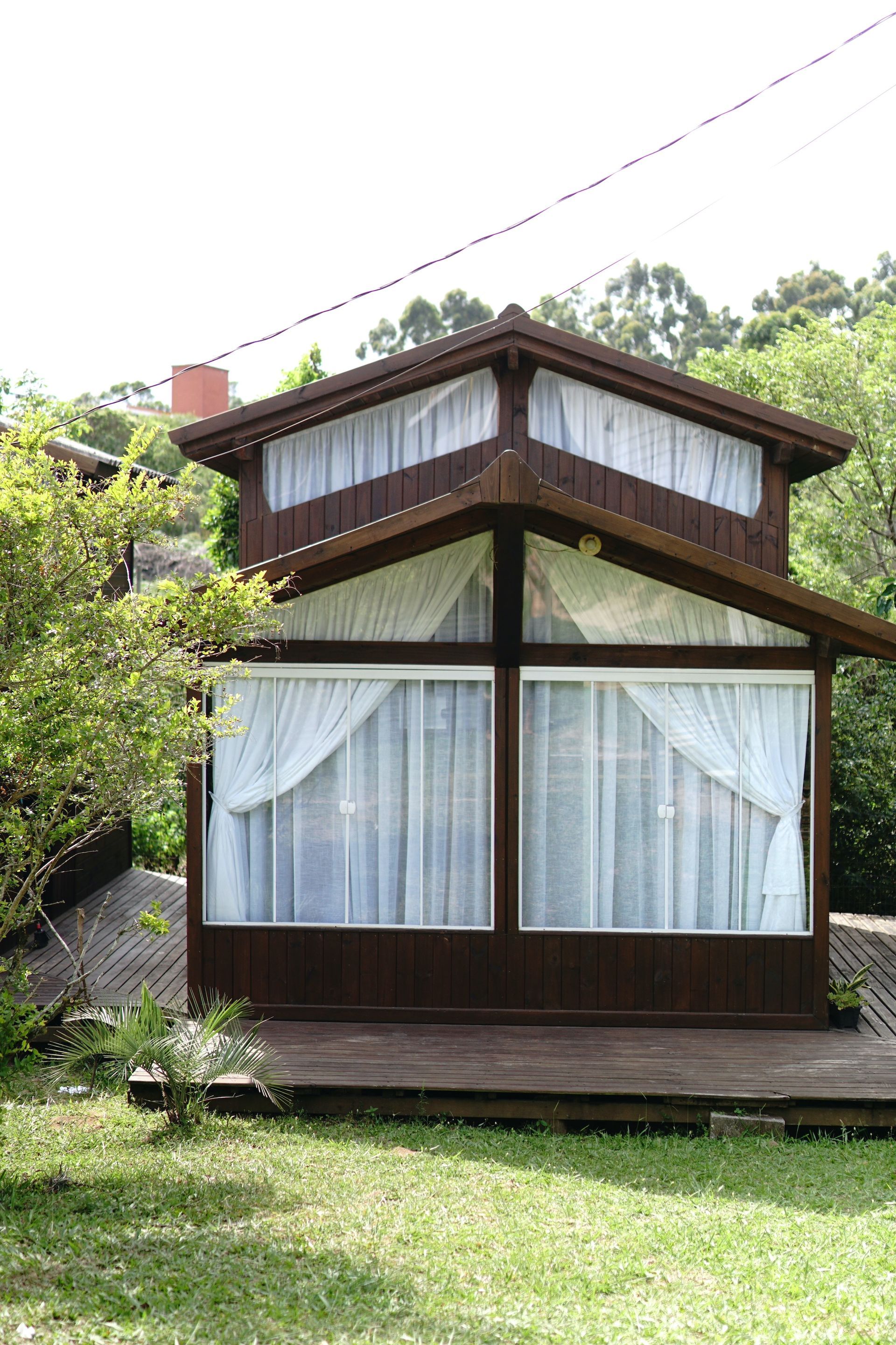 Pequena cabana de madeira com grandes janelas e cortinas brancas em uma encosta gramada, cercada por árvores.