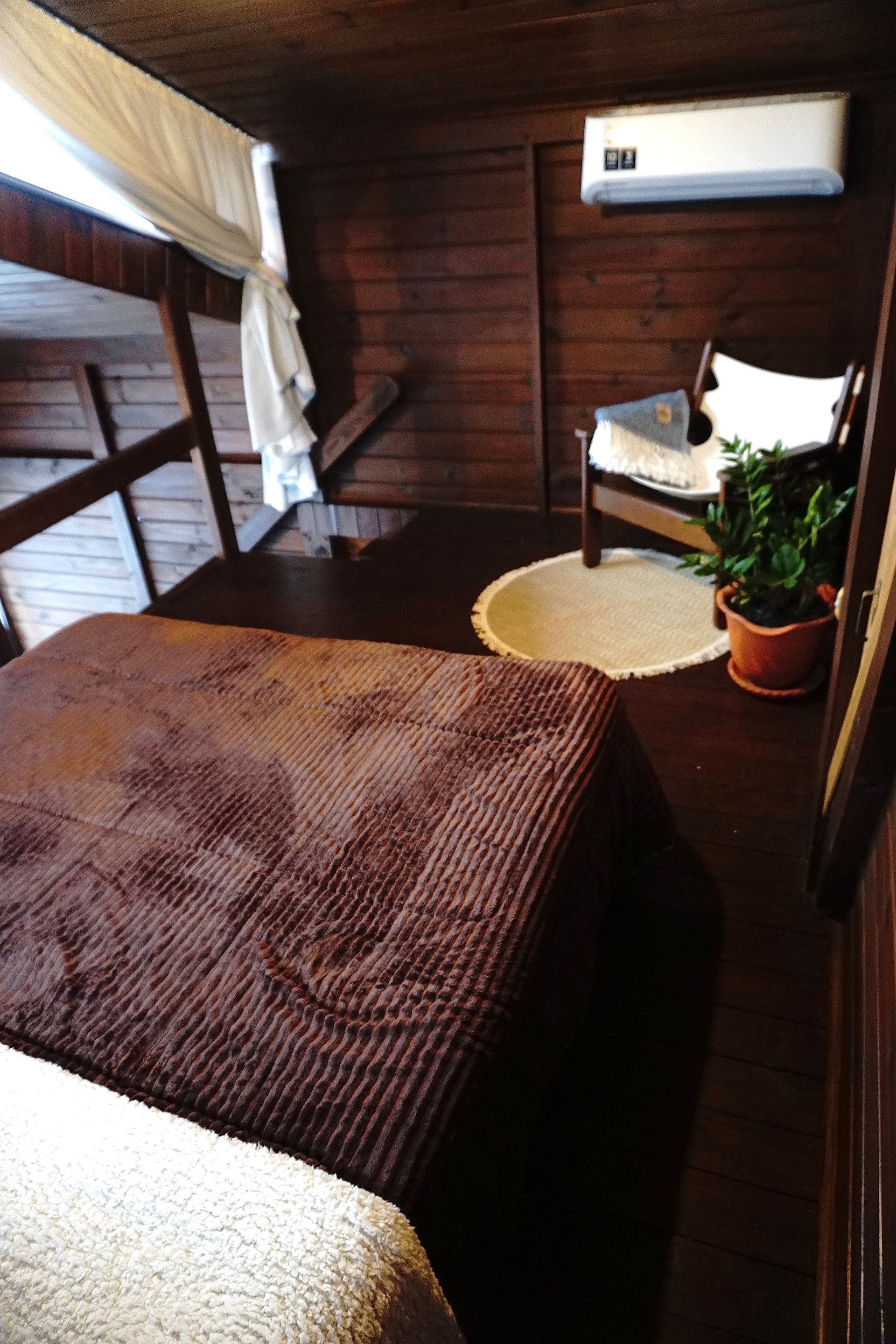 Quarto compacto com painéis de madeira, beliche, colcha marrom, escrivaninha, cadeira e ar-condicionado de parede.