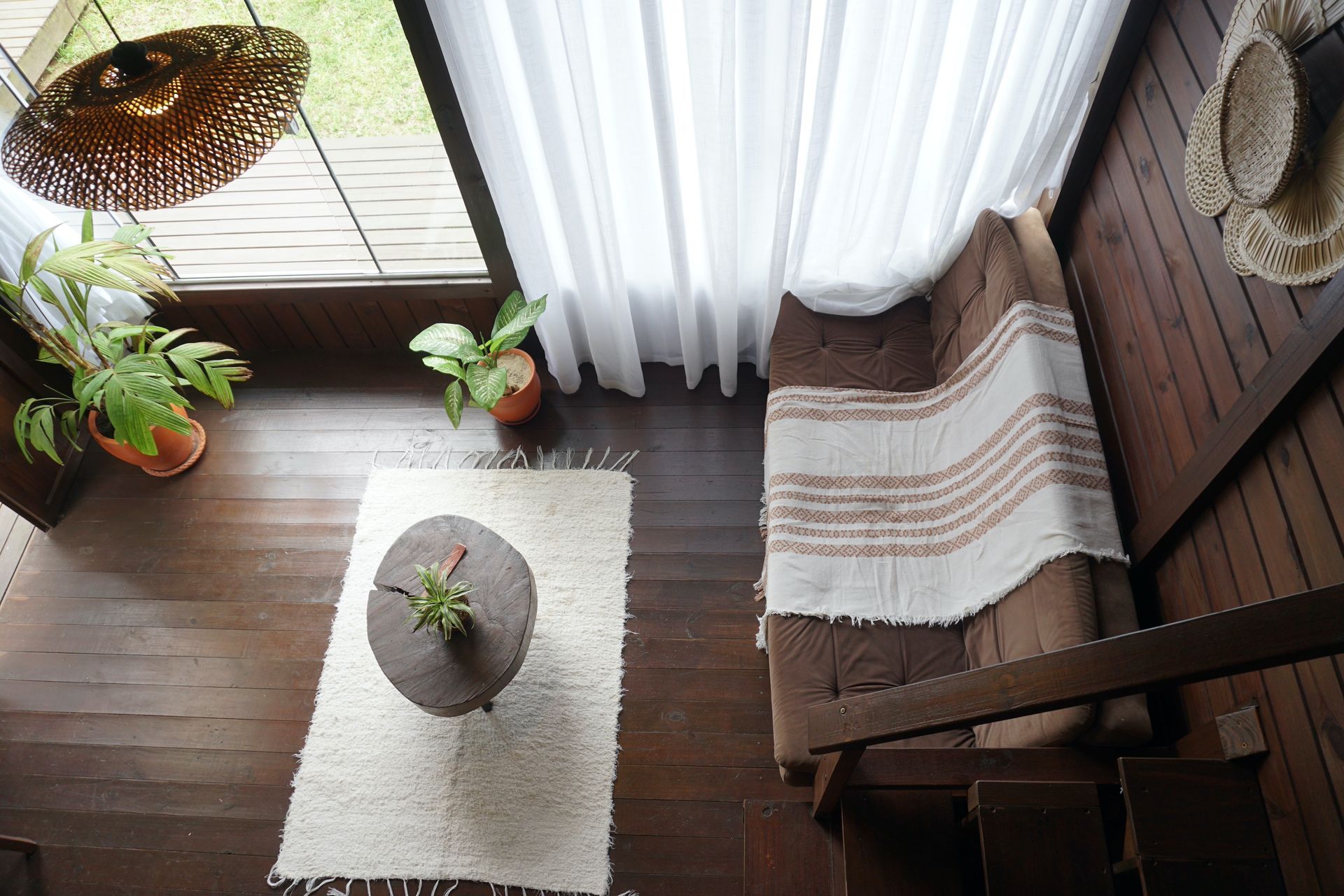 Sala de estar aconchegante e ensolarada com piso de madeira, tapete branco, plantas em vasos e uma poltrona listrada junto a cortinas translúcidas.
