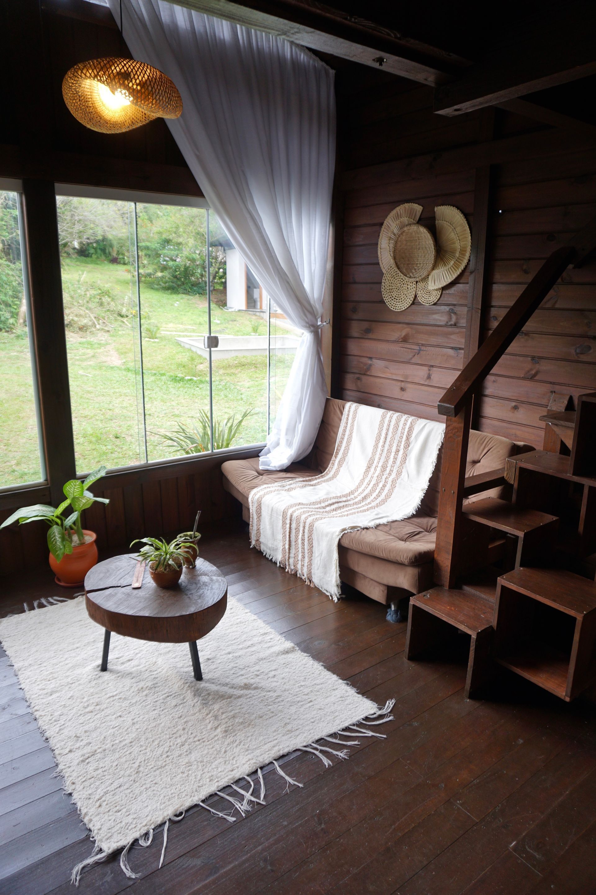 Sala de estar aconchegante em estilo rústico com uma cadeira branca, mesa de centro, tapete, luminária pendente e uma grande janela com cortinas translúcidas.