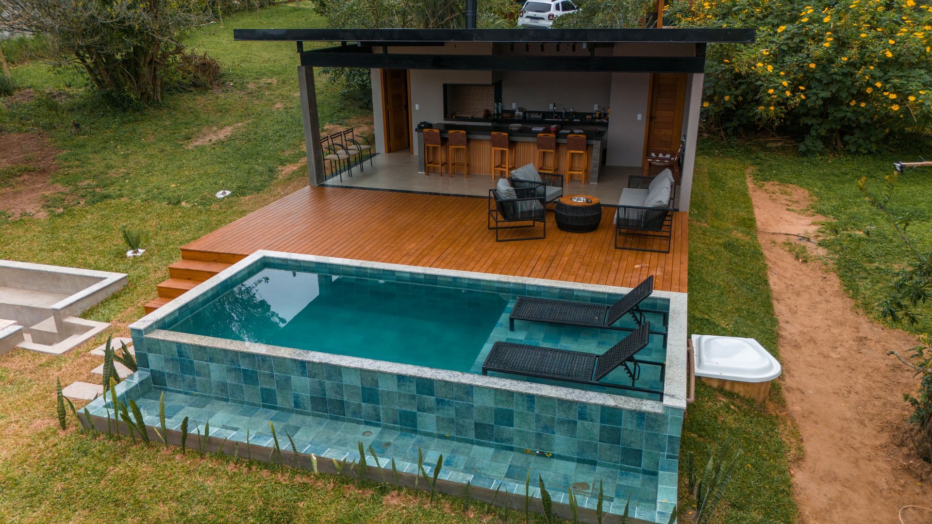 Pequena piscina no quintal ao lado de um pátio coberto com espreguiçadeiras e uma mesa de jantar com vista para uma encosta verdejante.
