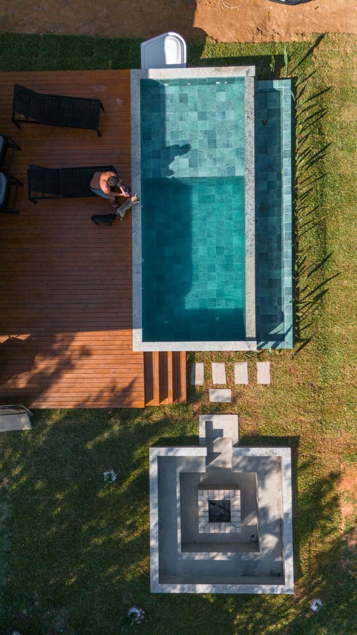 Vista aérea de uma piscina no quintal ao lado de um deck de madeira com espreguiçadeira e uma área quadrada com lareira.
