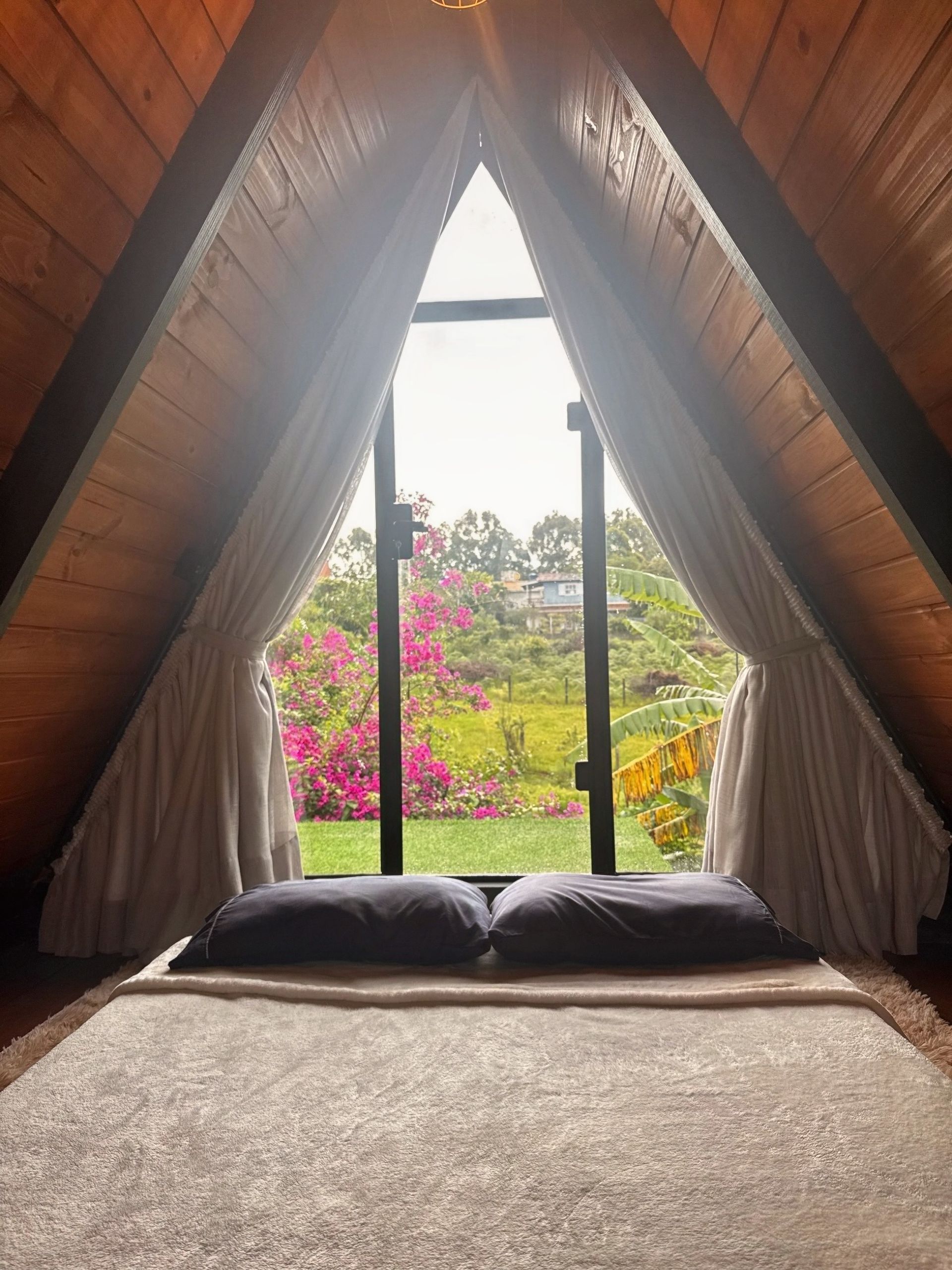 Janela em forma de A com cortinas, com vista para um jardim e um assento almofadado em primeiro plano.