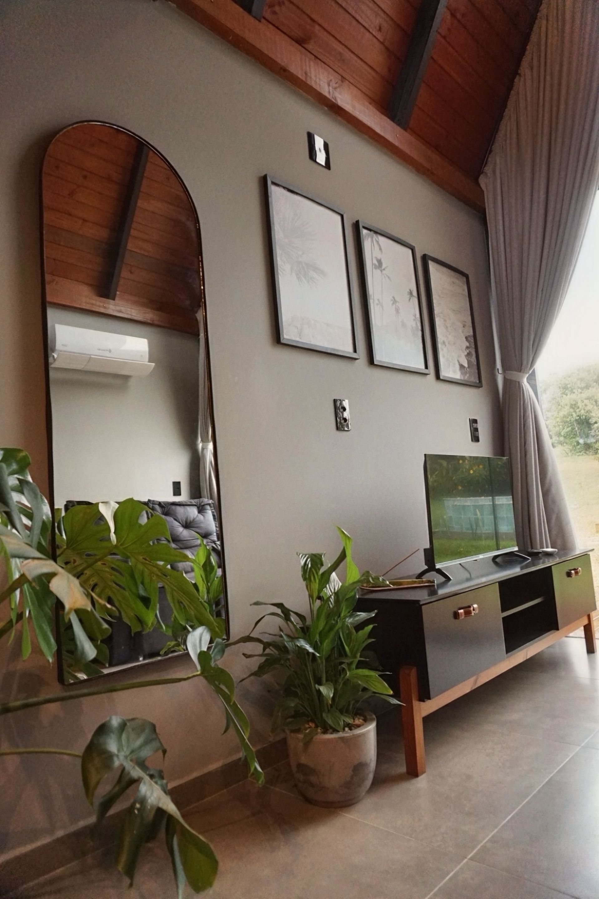 Sala de estar aconchegante com espelho grande, plantas em vasos, obras de arte na parede, suporte para TV e teto de madeira inclinado.
