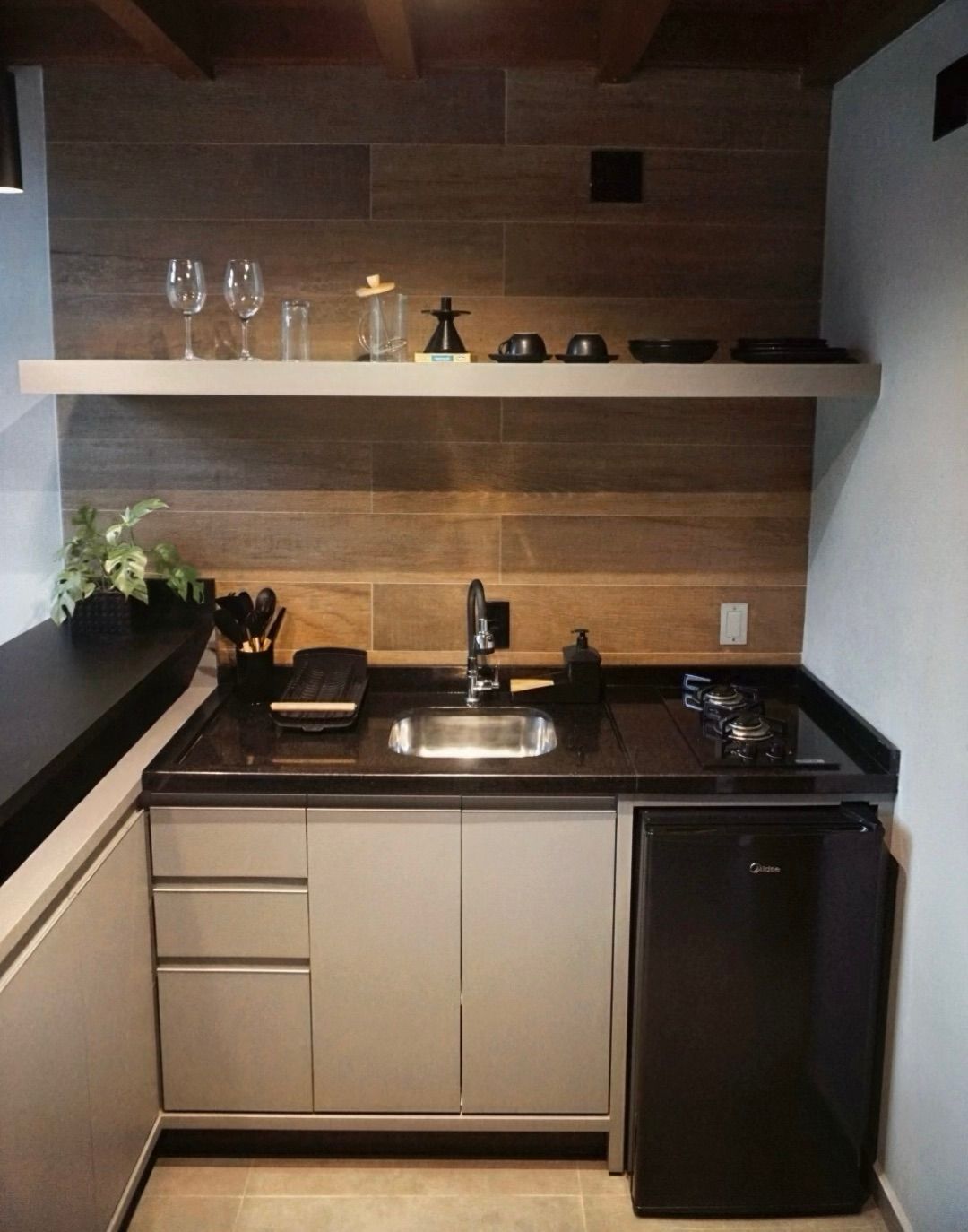 Cozinha compacta e moderna com painel de madeira na parede, bancada preta, pia, fogão, frigobar e prateleira aberta.