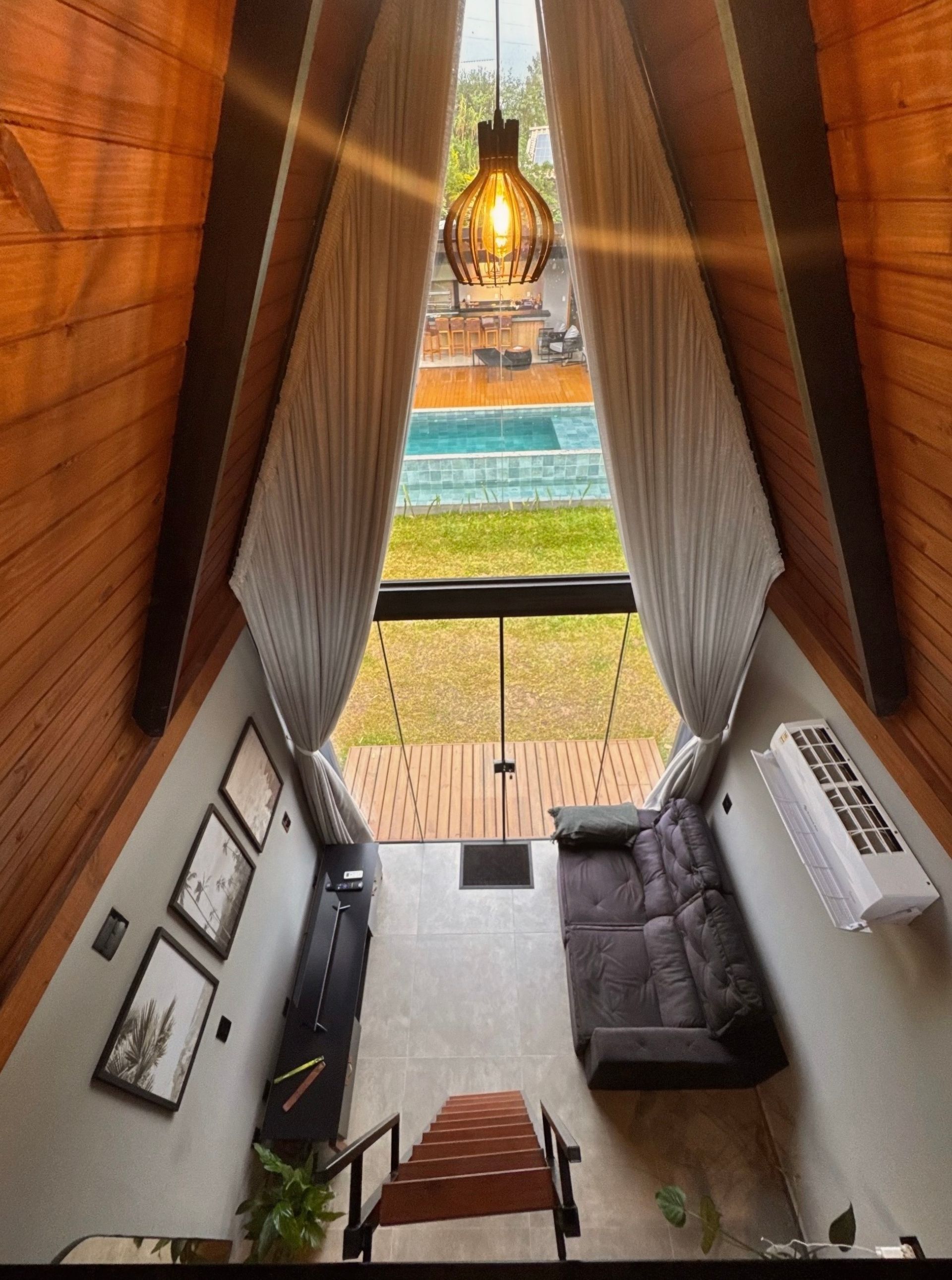 Uma sala de estar triangular com um candeeiro suspenso, sofá cinzento, paredes de madeira e vista para a piscina e para o oceano a partir da varanda.