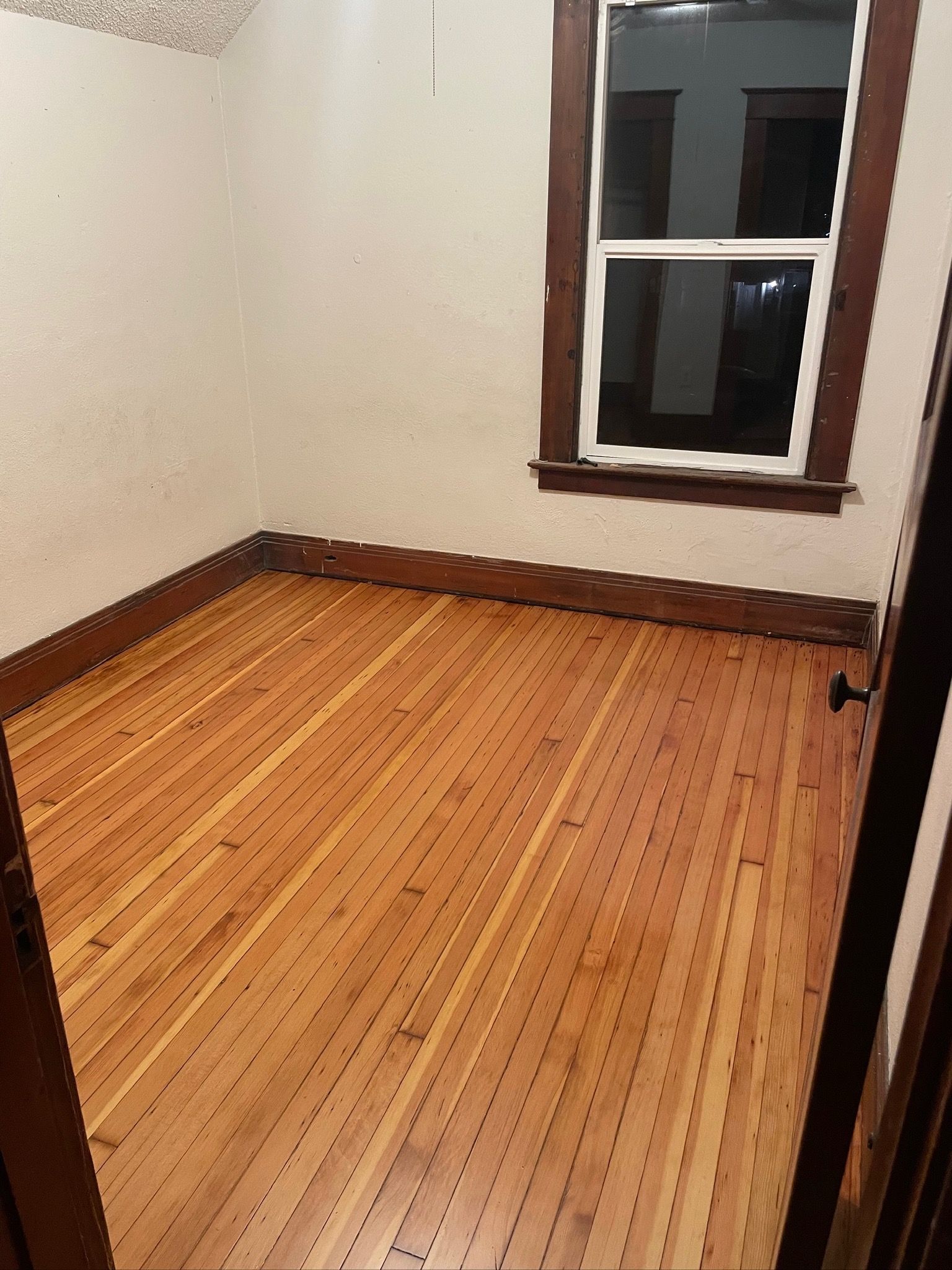 A small room with light wood floors, brown baseboards, and a window with a dark wooden frame.