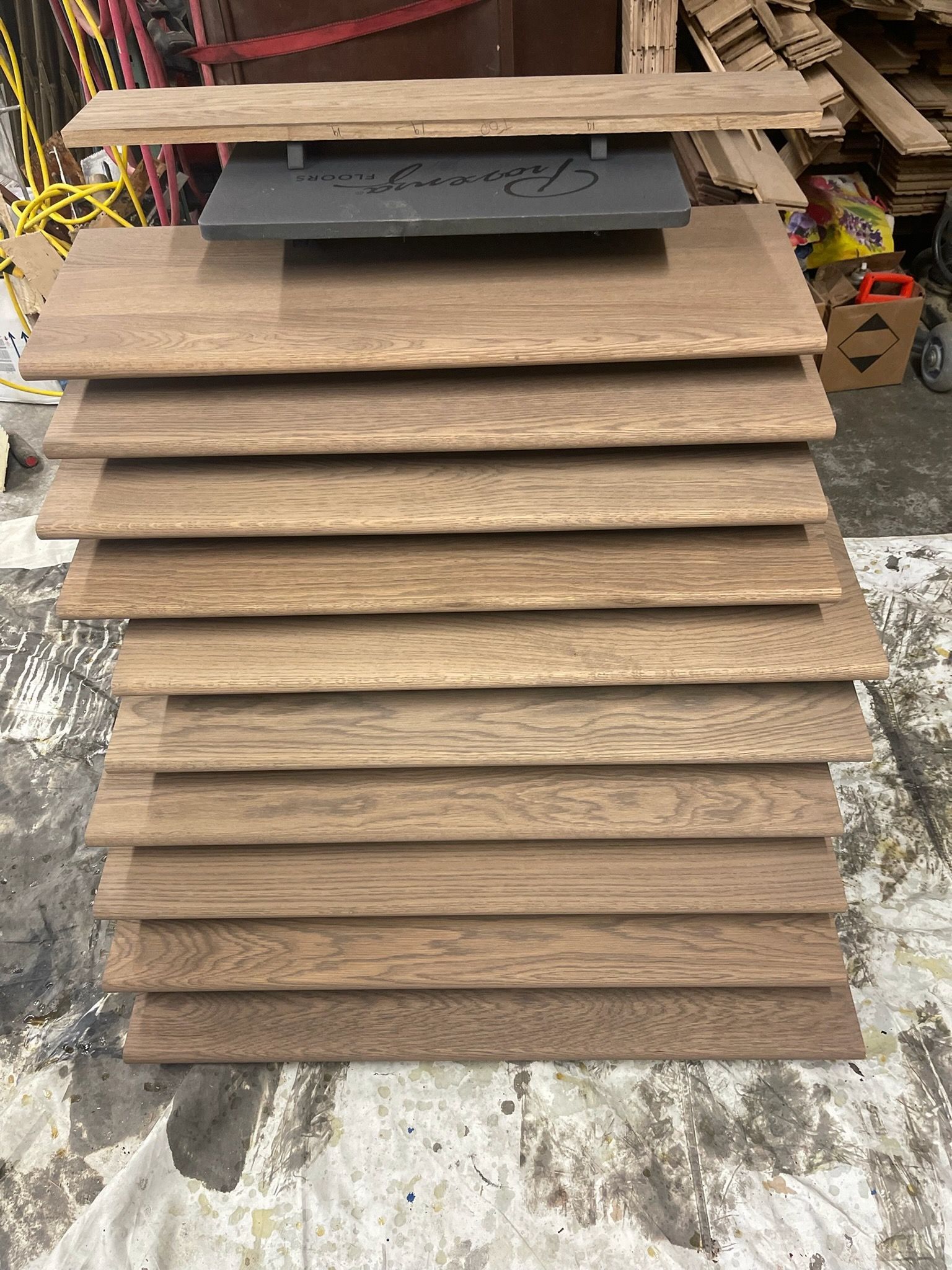 A vertical stack of twelve light brown wooden stair treads sits on a concrete floor in a workshop.