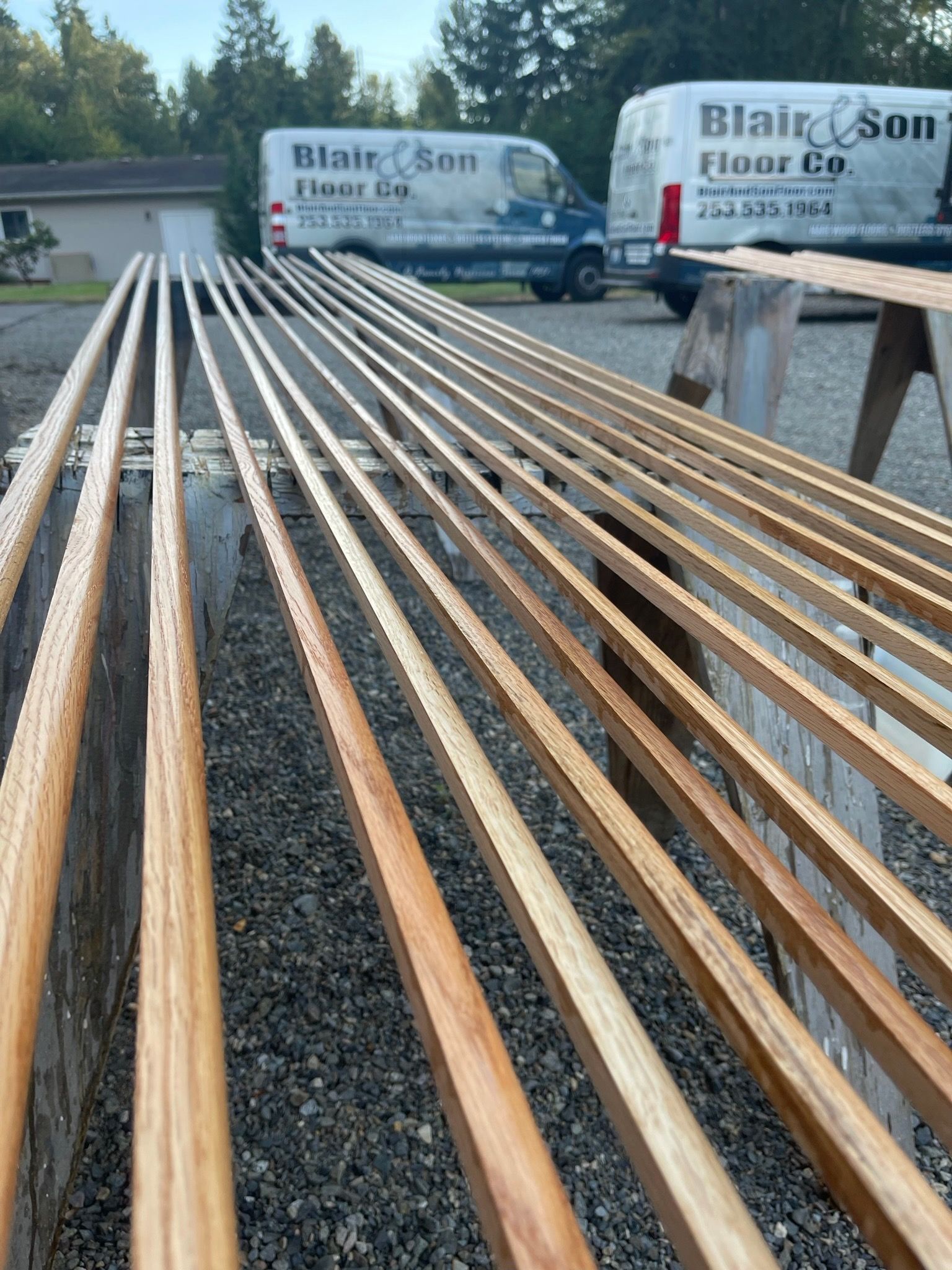 Several long, finished wooden floor trim pieces rest on sawhorses outdoors, with two company vans in the background.