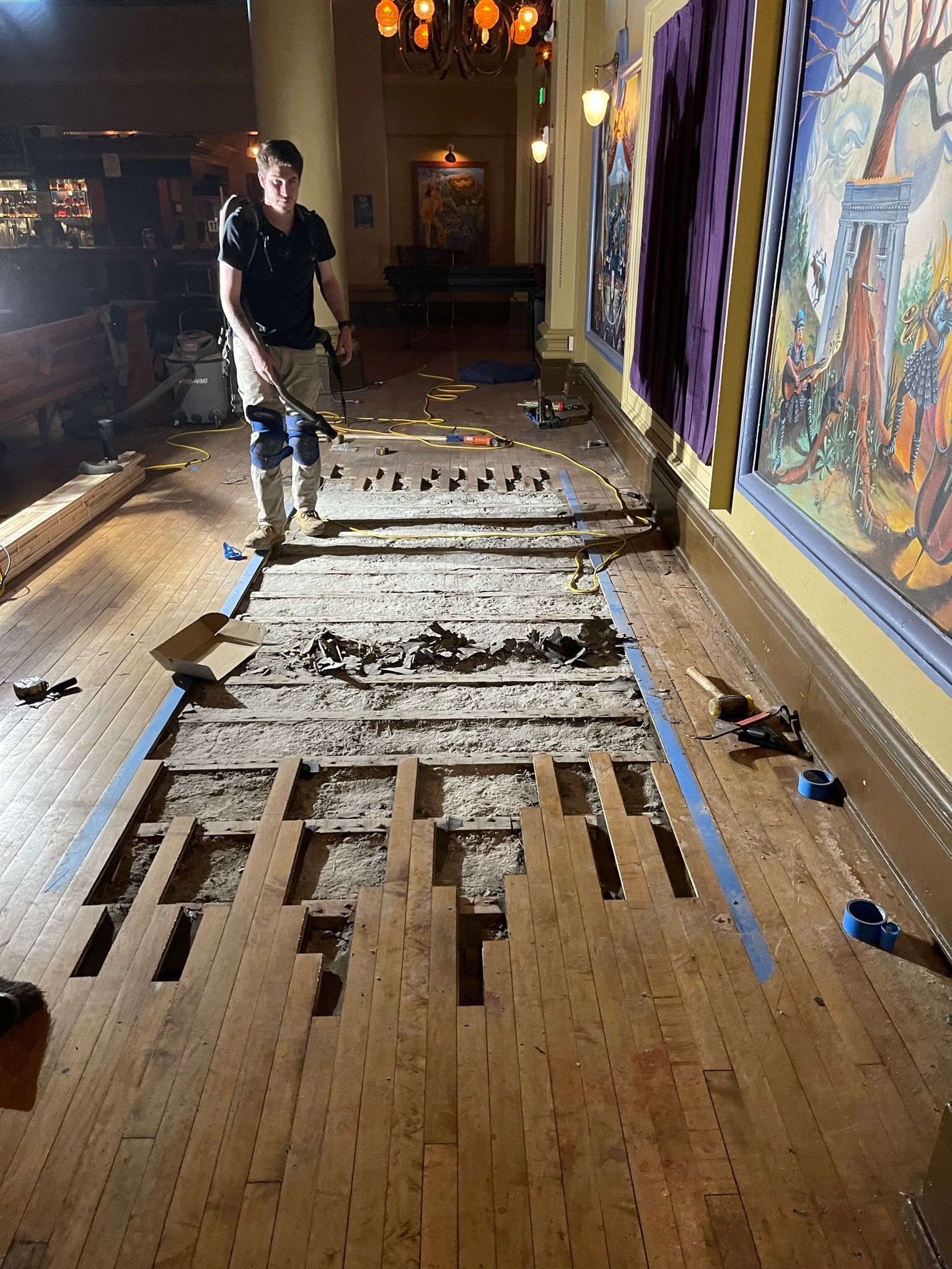 A person stands in a hallway during wooden floor renovation, with boards removed to expose the subfloor below.