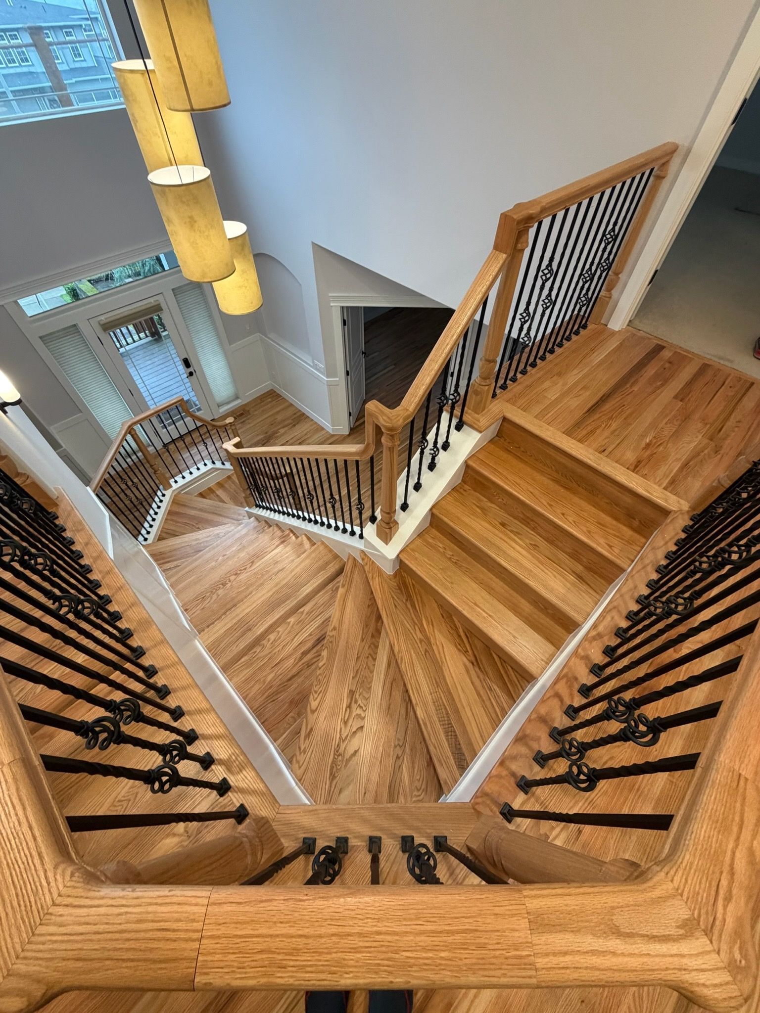A high-angle view of a wooden staircase with black iron balusters, descending toward a light-filled entryway.