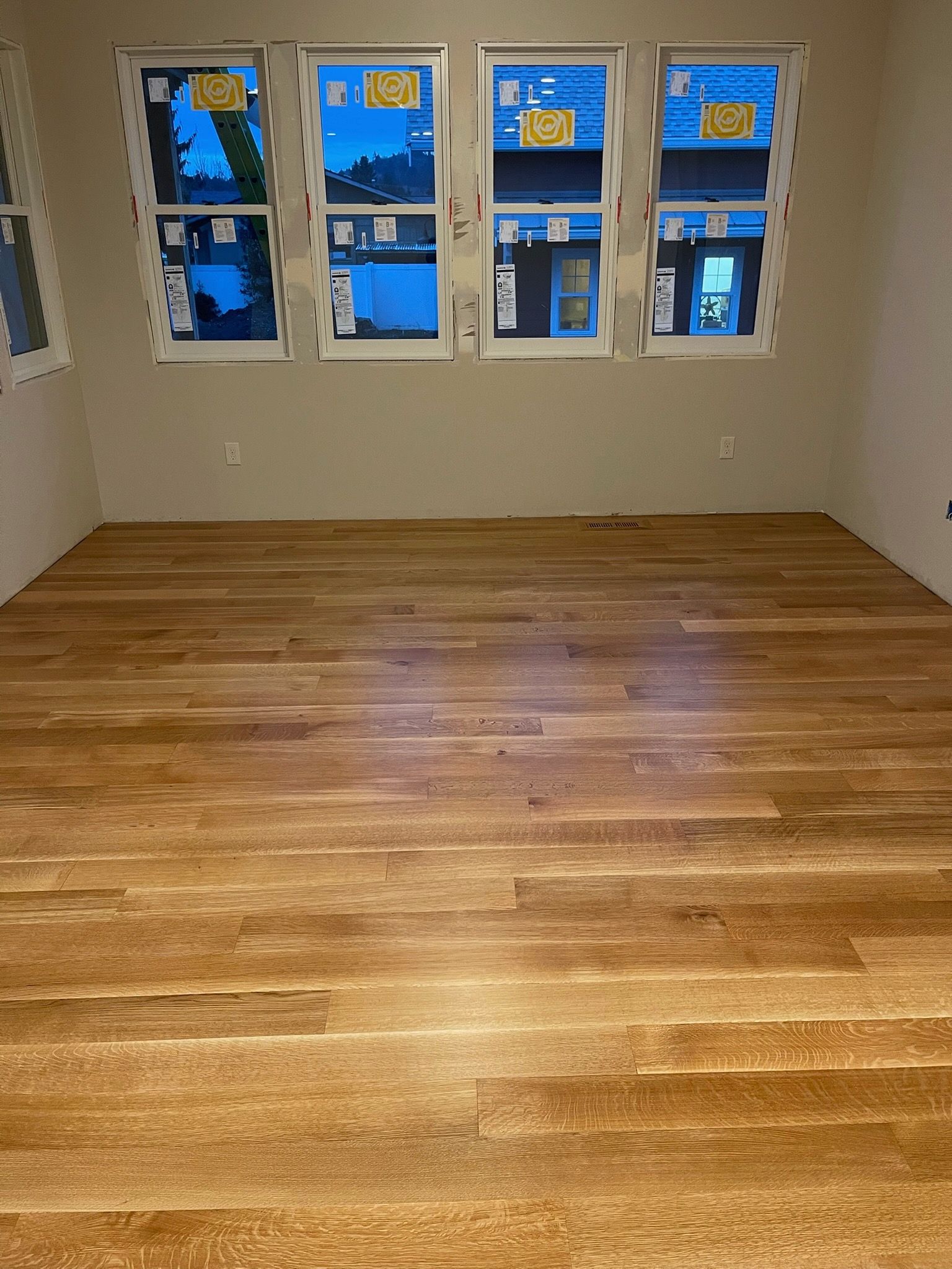 A room with light-colored hardwood floors and four windows featuring protective film, set against plain, neutral walls.