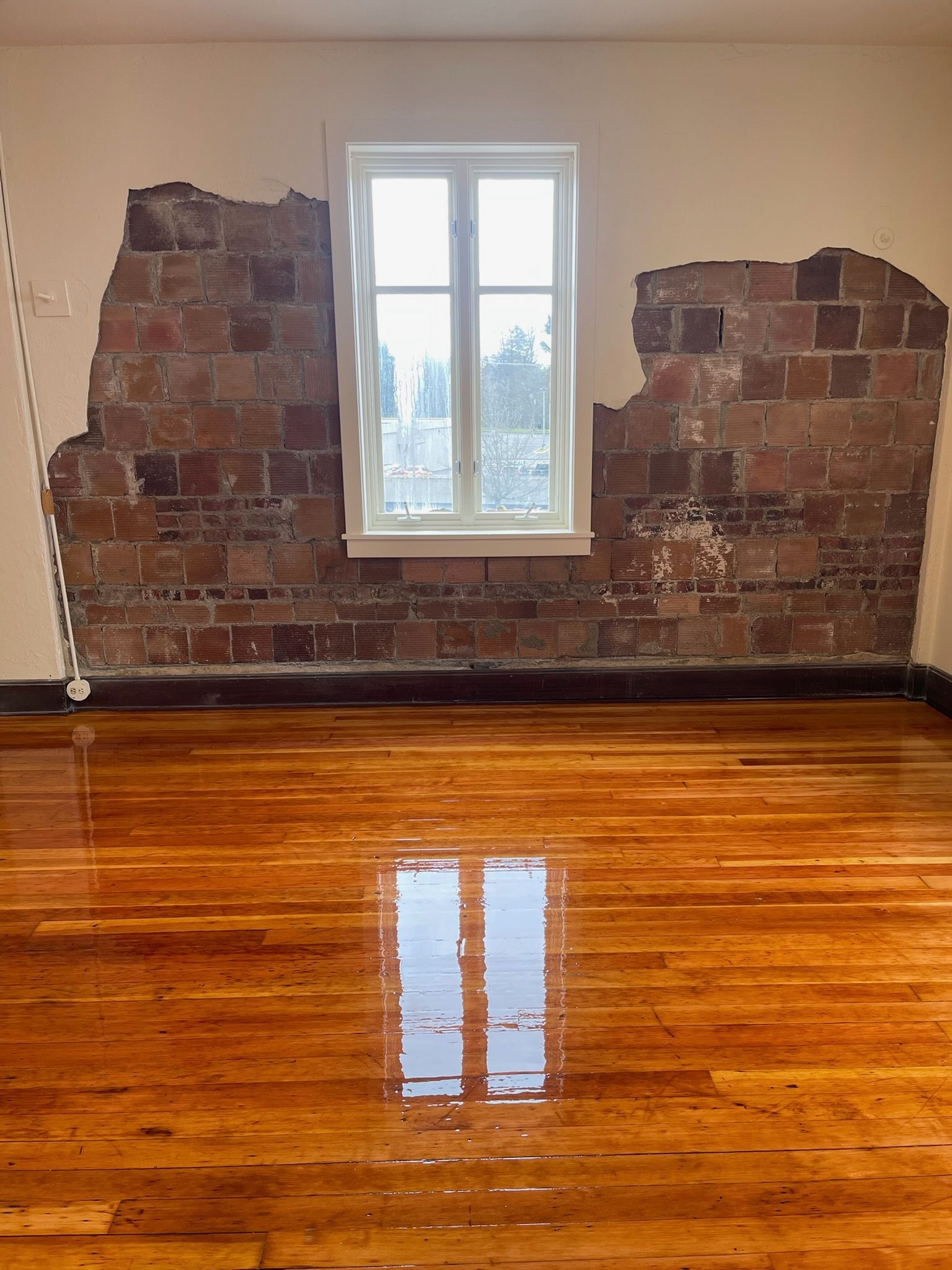 A room with a polished wood floor and a wall where plaster is partially removed, revealing red brick underneath.