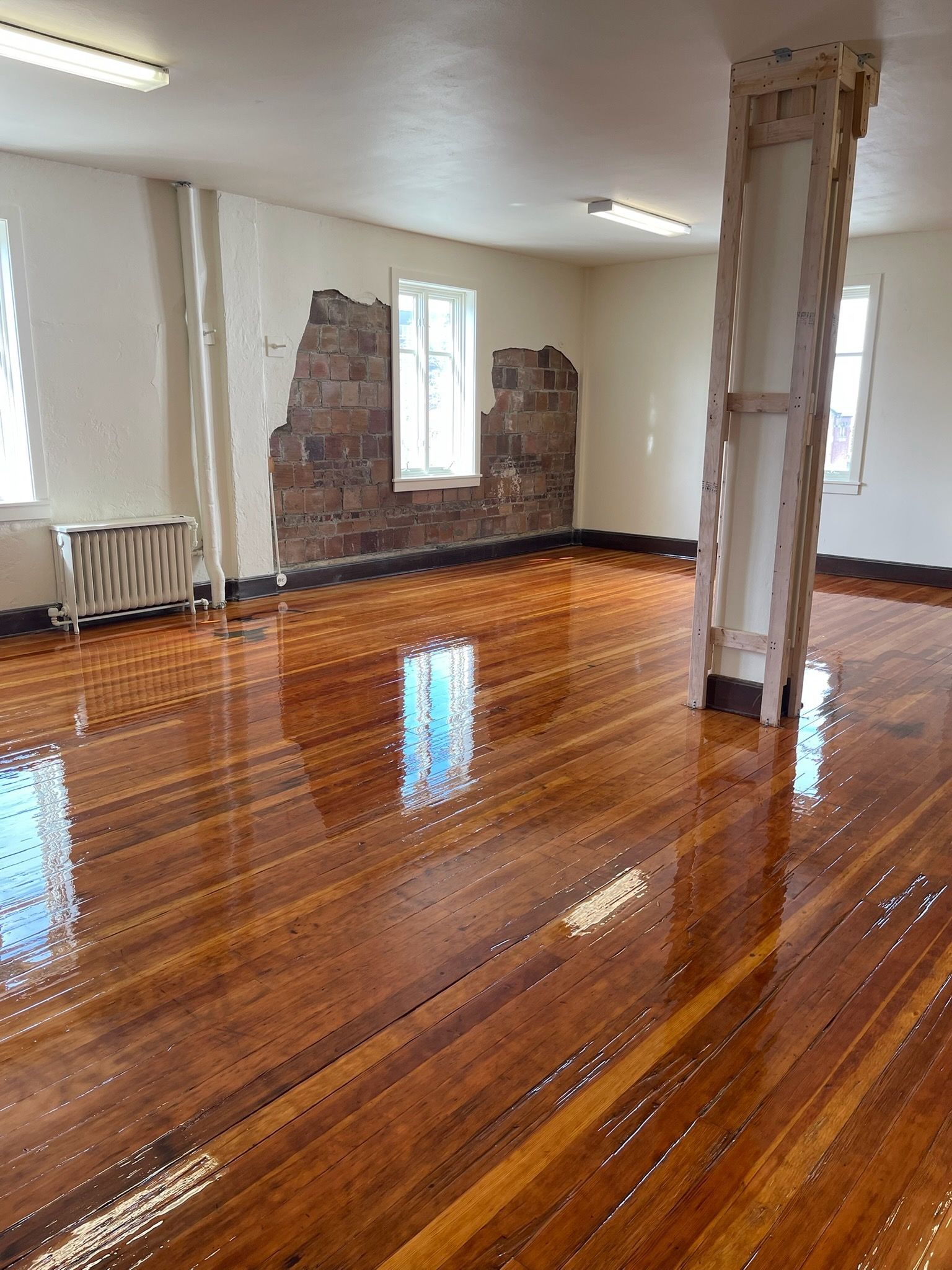 Empty room with polished hardwood floors, two windows, a central support column, and a section of exposed brick wall.