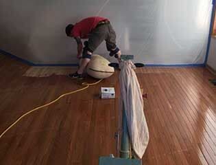 A person in a red shirt works on hardwood flooring in a room covered with plastic sheeting.
