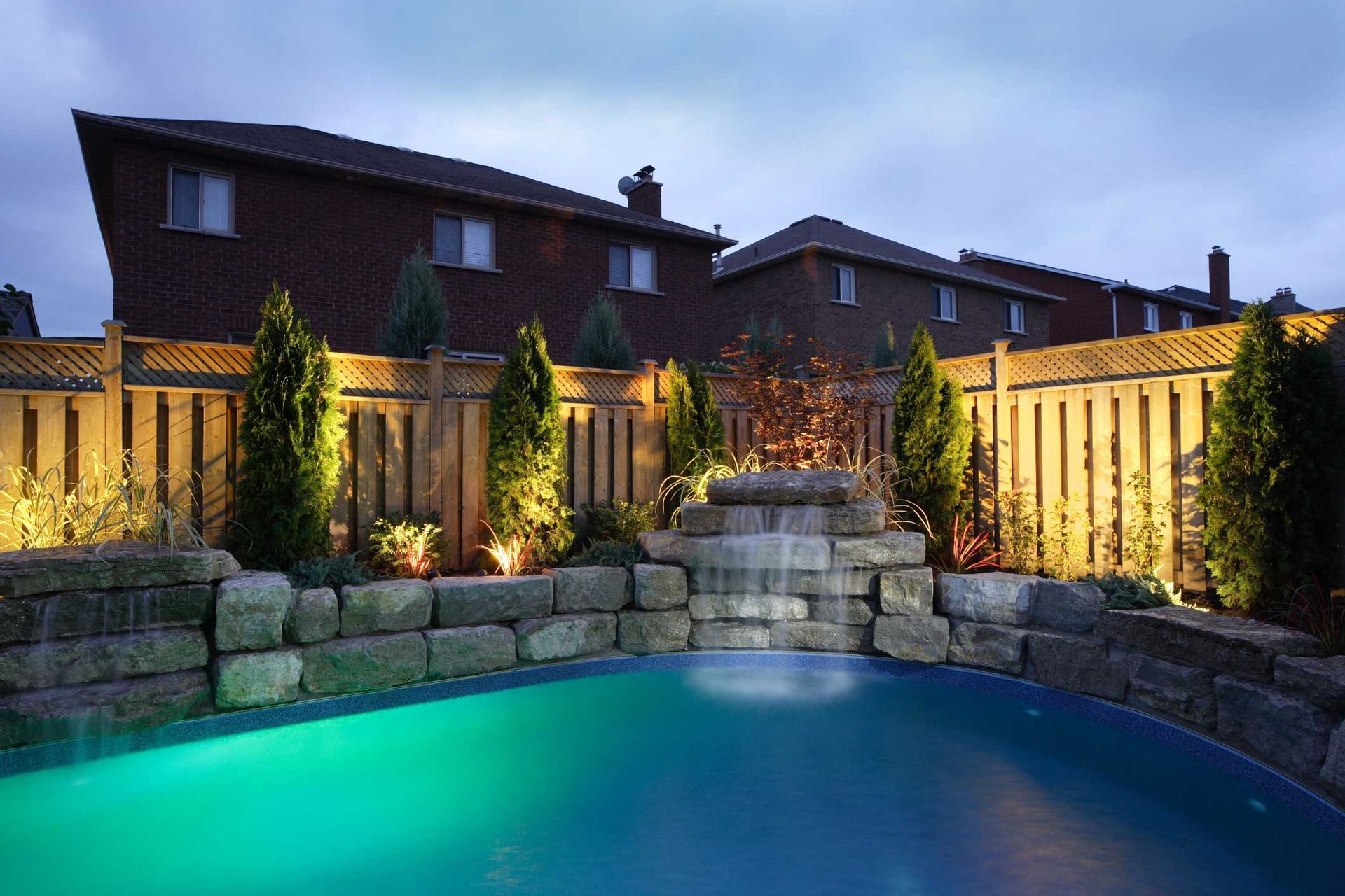 A swimming pool with a waterfall in the backyard is lit up at night.