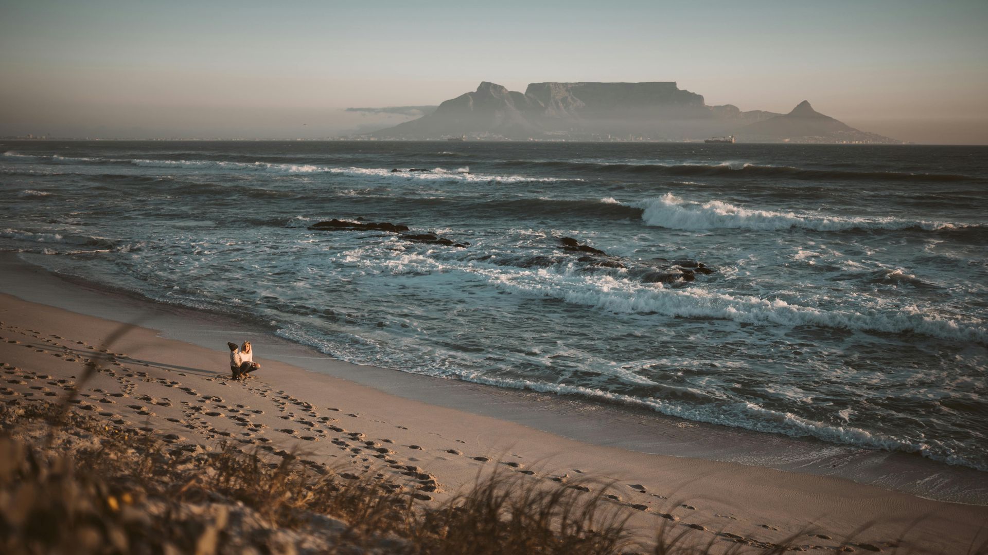 A person is walking on a beach near the ocean with a mountain in the background.
