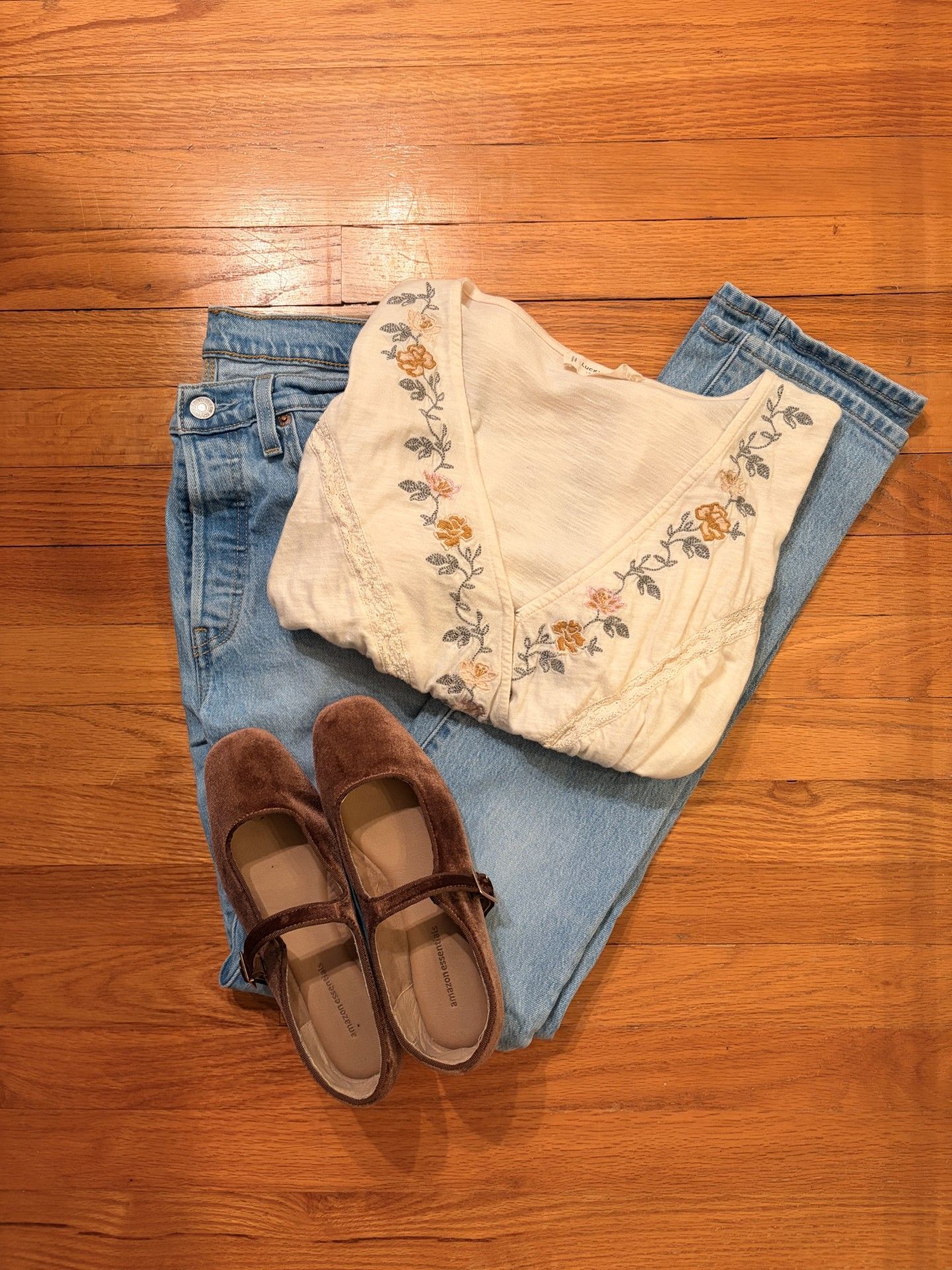 Blue jeans, cream embroidered blouse, and brown sandals arranged on a wooden floor.