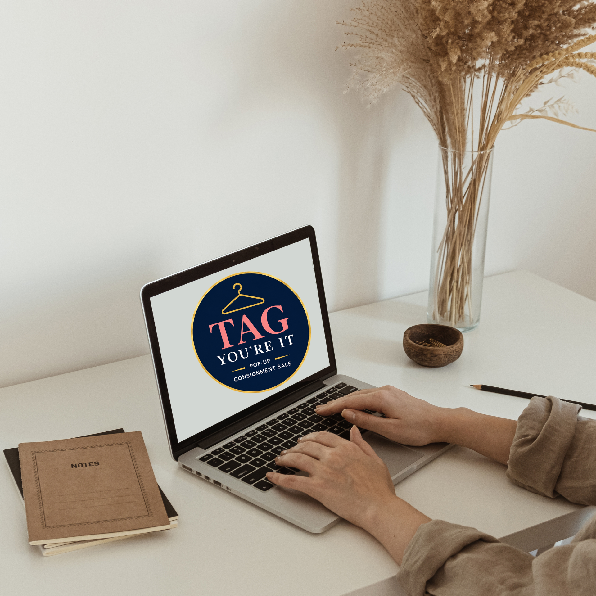 Hands typing on a laptop at a white desk with a notebook and dried flowers; screen shows a logo.