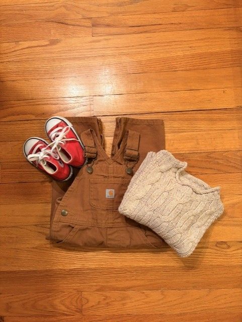 Children's outfit laid out on a wooden floor: brown overalls, red sneakers, and a cream knit sweater.