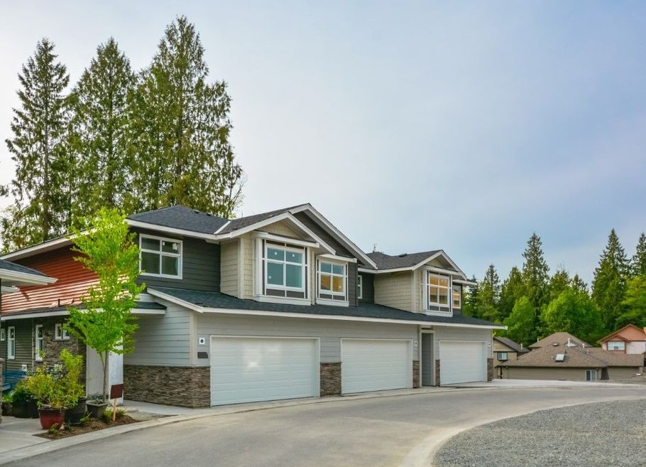 Suburban two-story gray house with white garage doors, stone accents, and tall trees in the yard