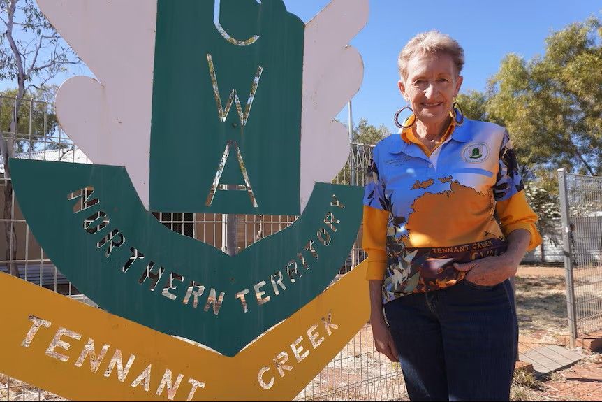 Person standing beside a green and gold sign reading “CWA” and “Tennant Creek” outdoors