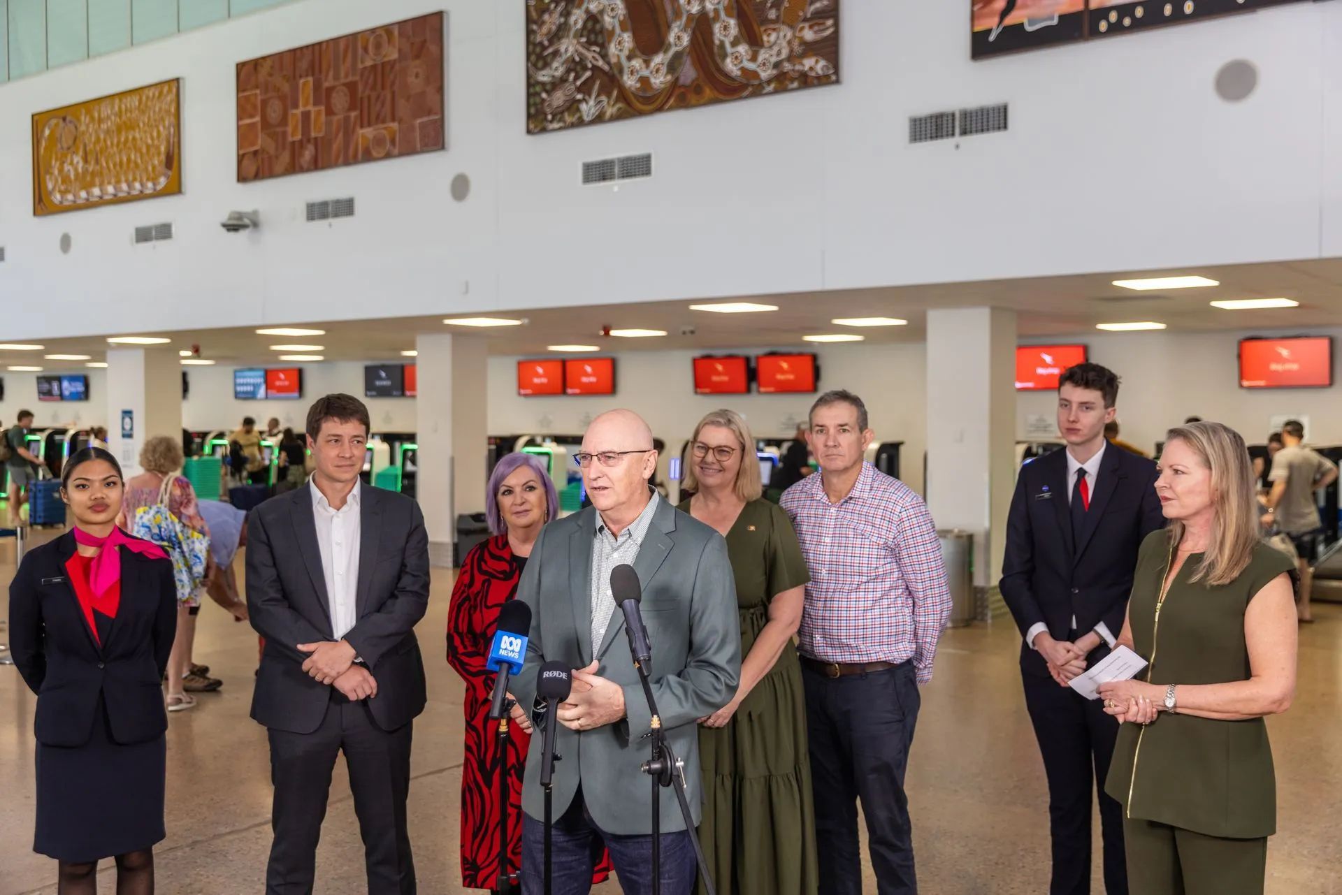 Group of people at an airport, one speaking into a microphone. White walls, art, and flight information boards are visible.