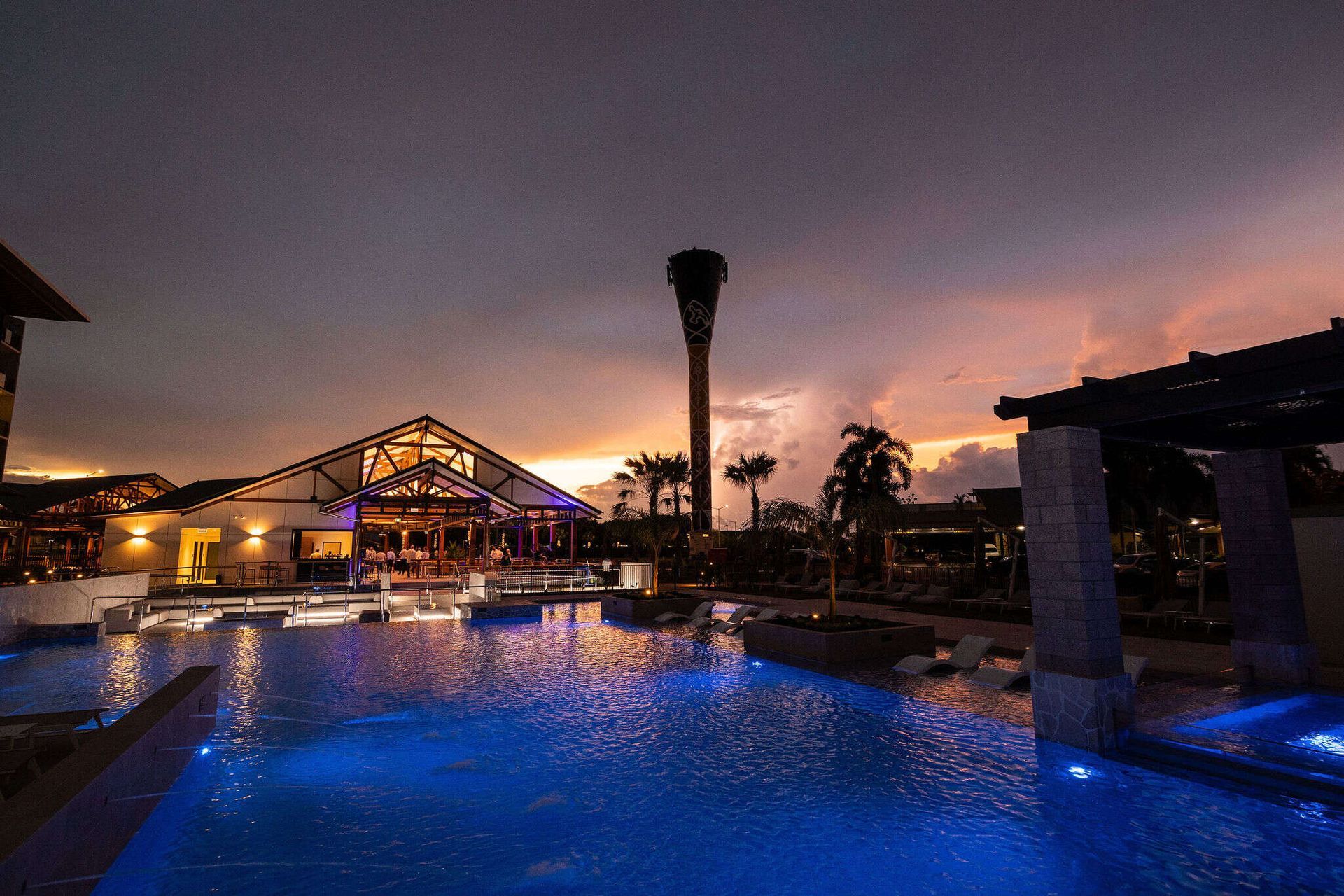 A large swimming pool is lit up at night with palm trees in the background