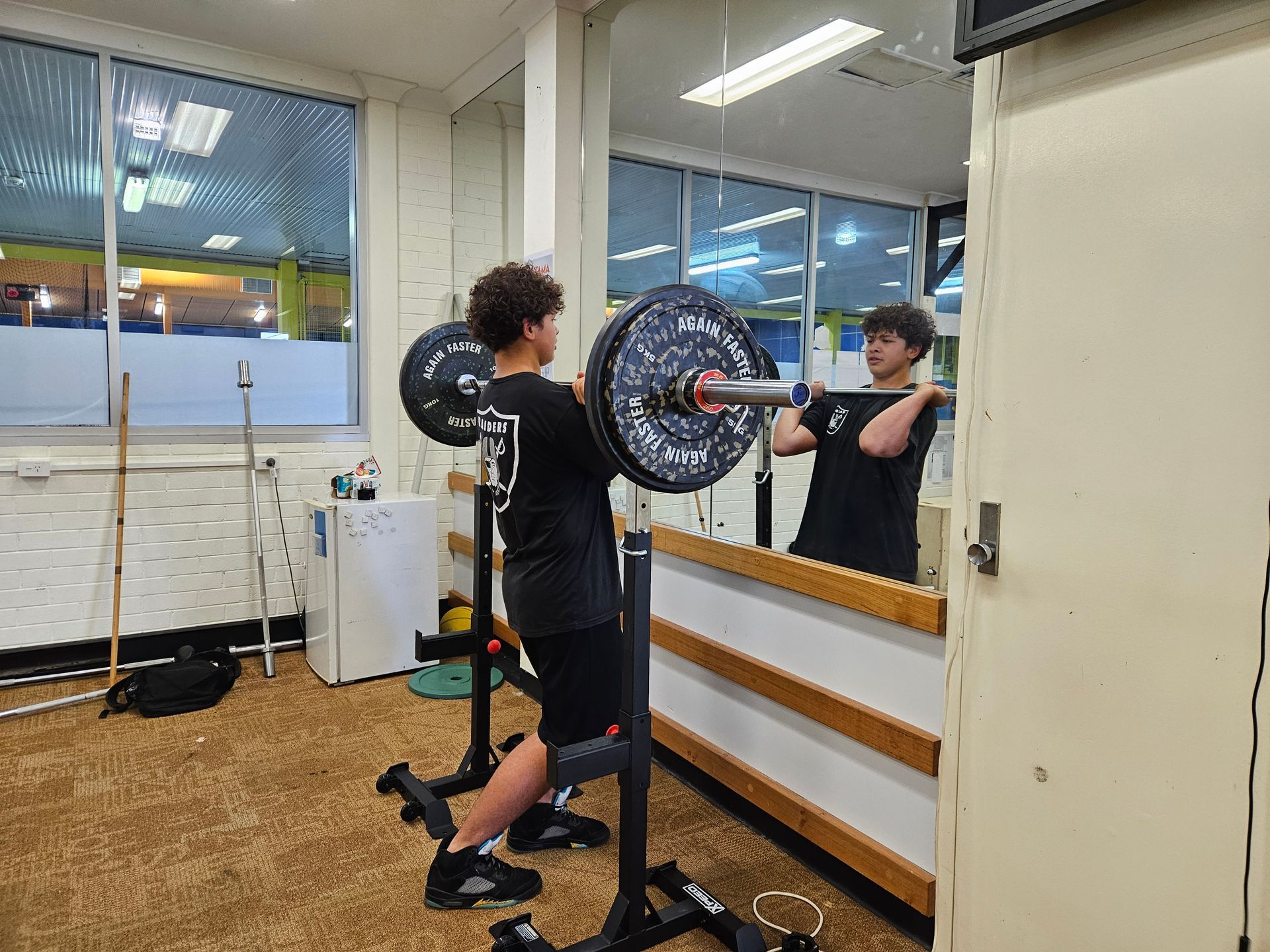 Person squatting with a barbell in a gym, reflected in a mirror. They wear black clothing and sneakers.