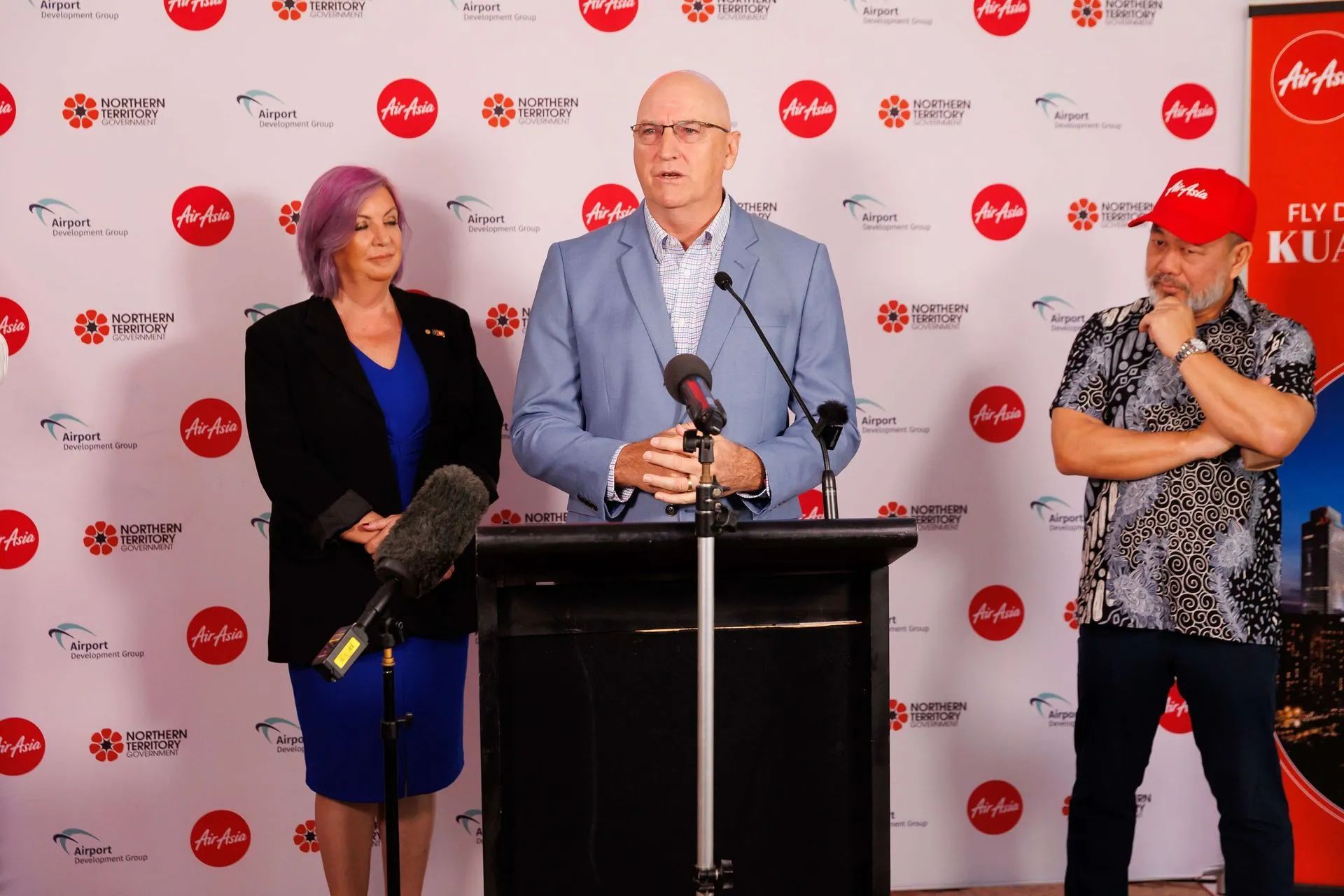 Three people at a podium, AirAsia event. Man speaks, flanked by woman with purple hair and man in red cap.