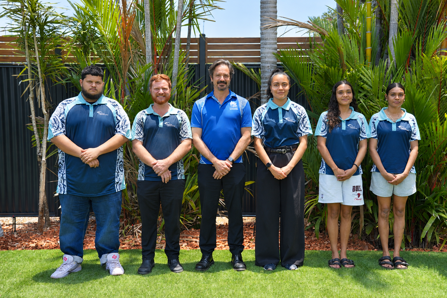 Six people standing on a lawn in blue uniform shirts in front of a building and palm trees