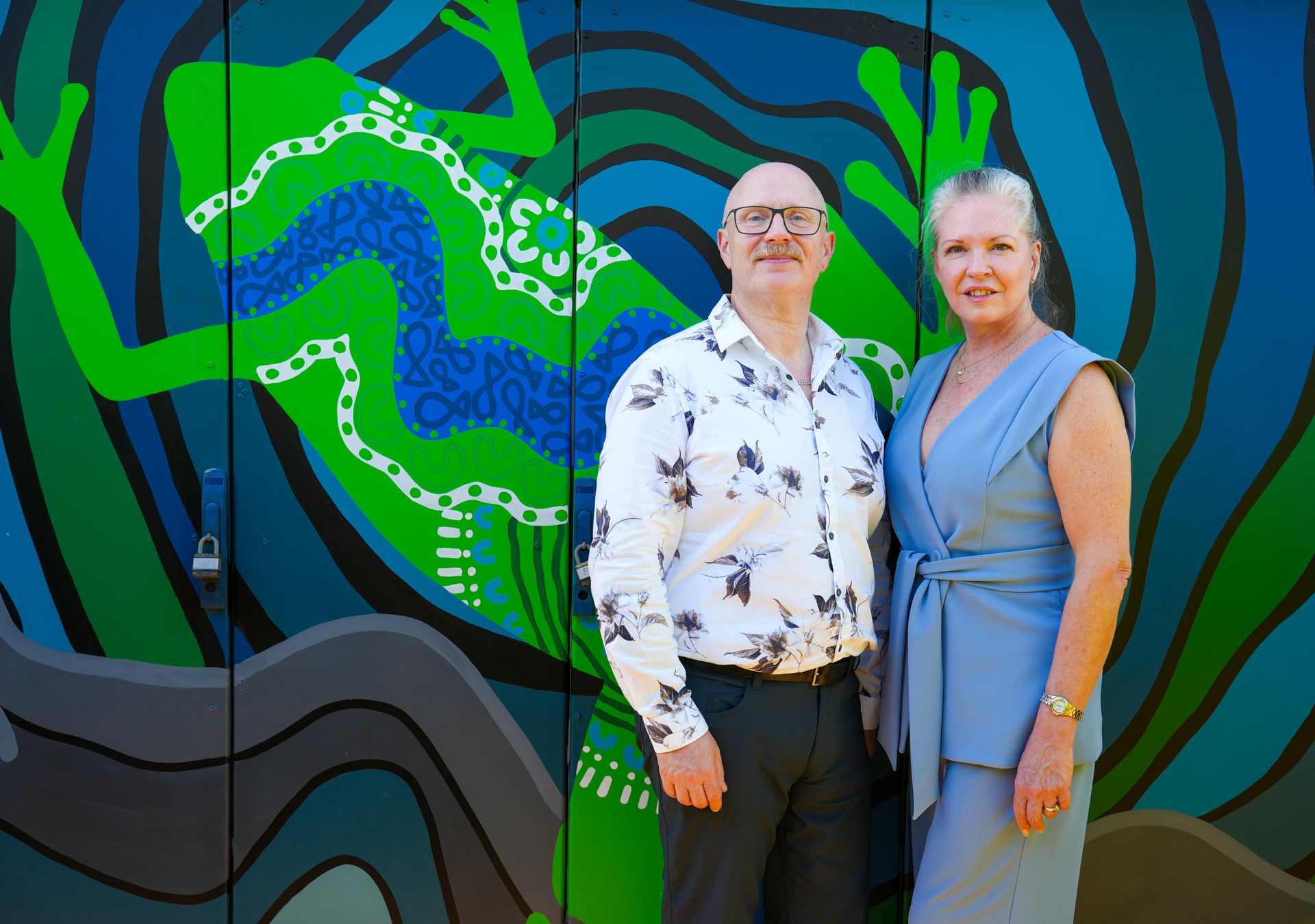 Two adults standing in front of a blue, green, and black mural, smiling at the camera.