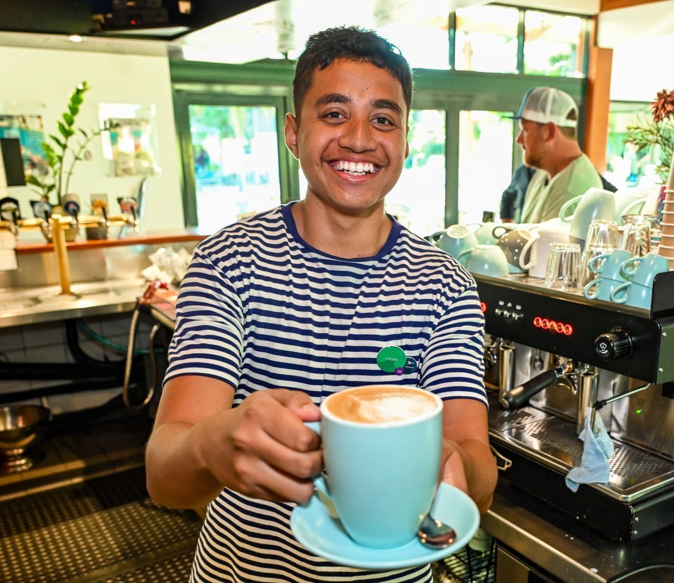 Young man with a big smile, holding out a light blue coffee cup. Cafe setting, wearing striped shirt.