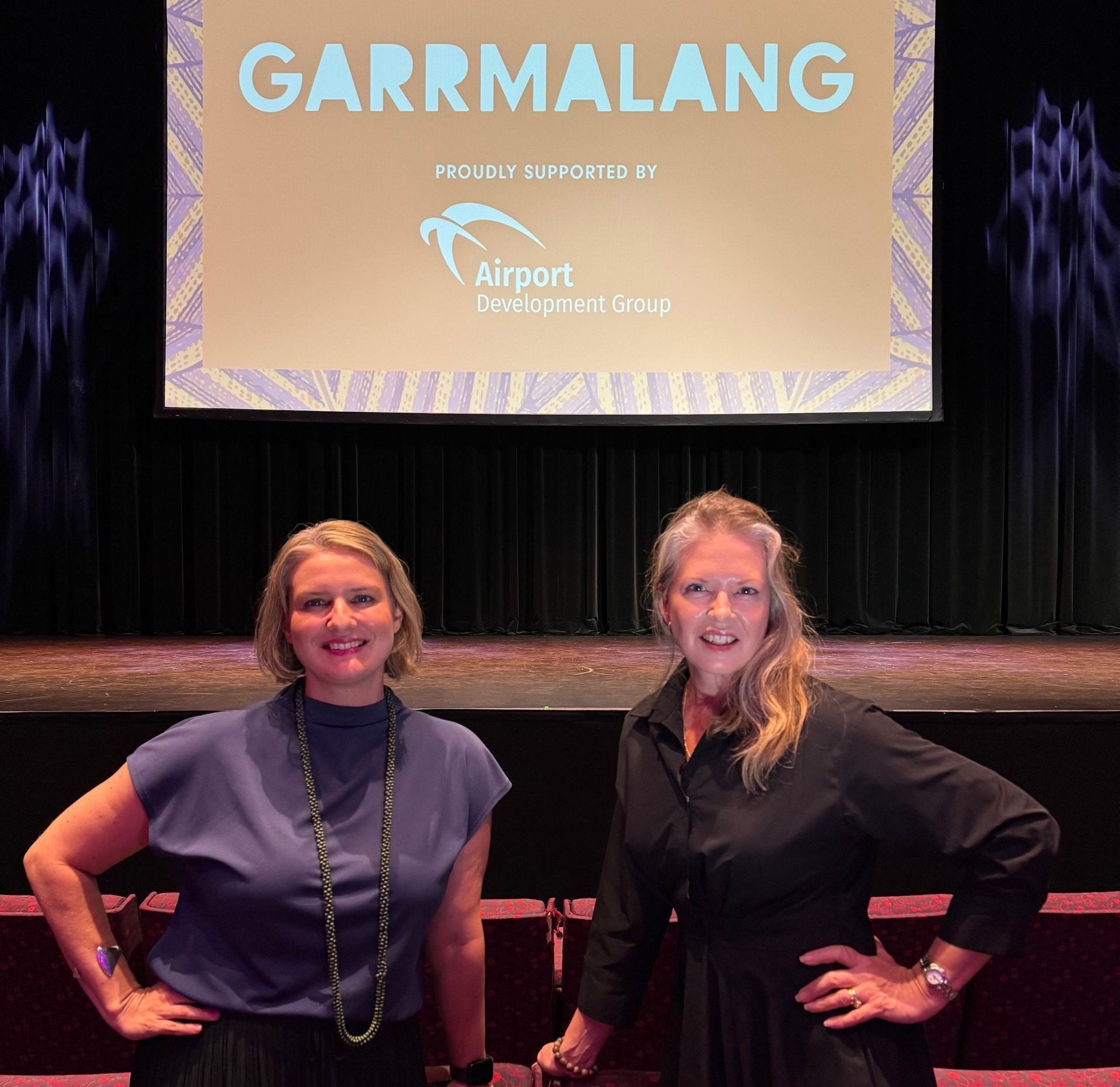 Two women standing in front of a stage, a screen with the word 