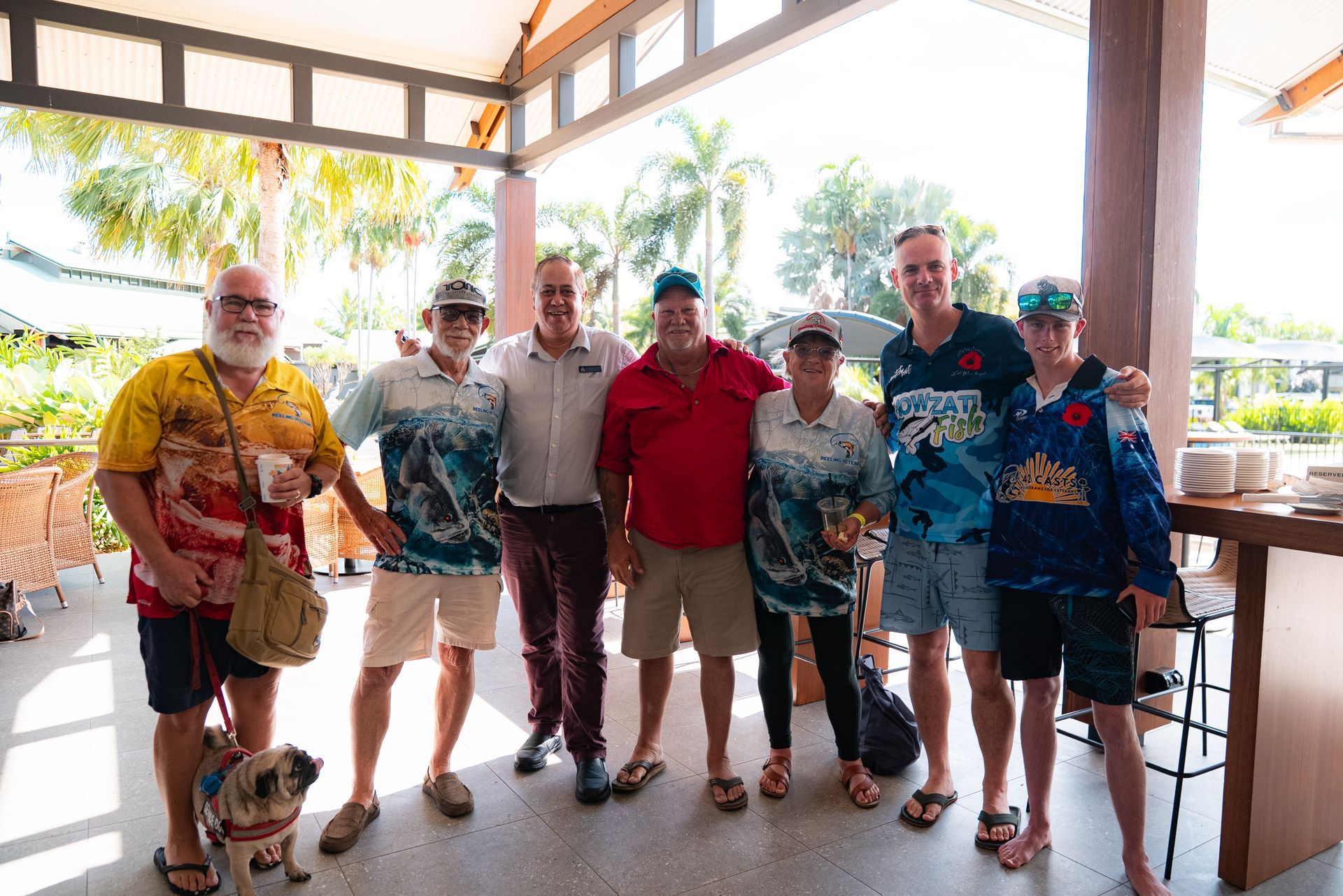 Group of people smiling, posing outdoors. Some wear tropical shirts, in a covered area with plants visible.