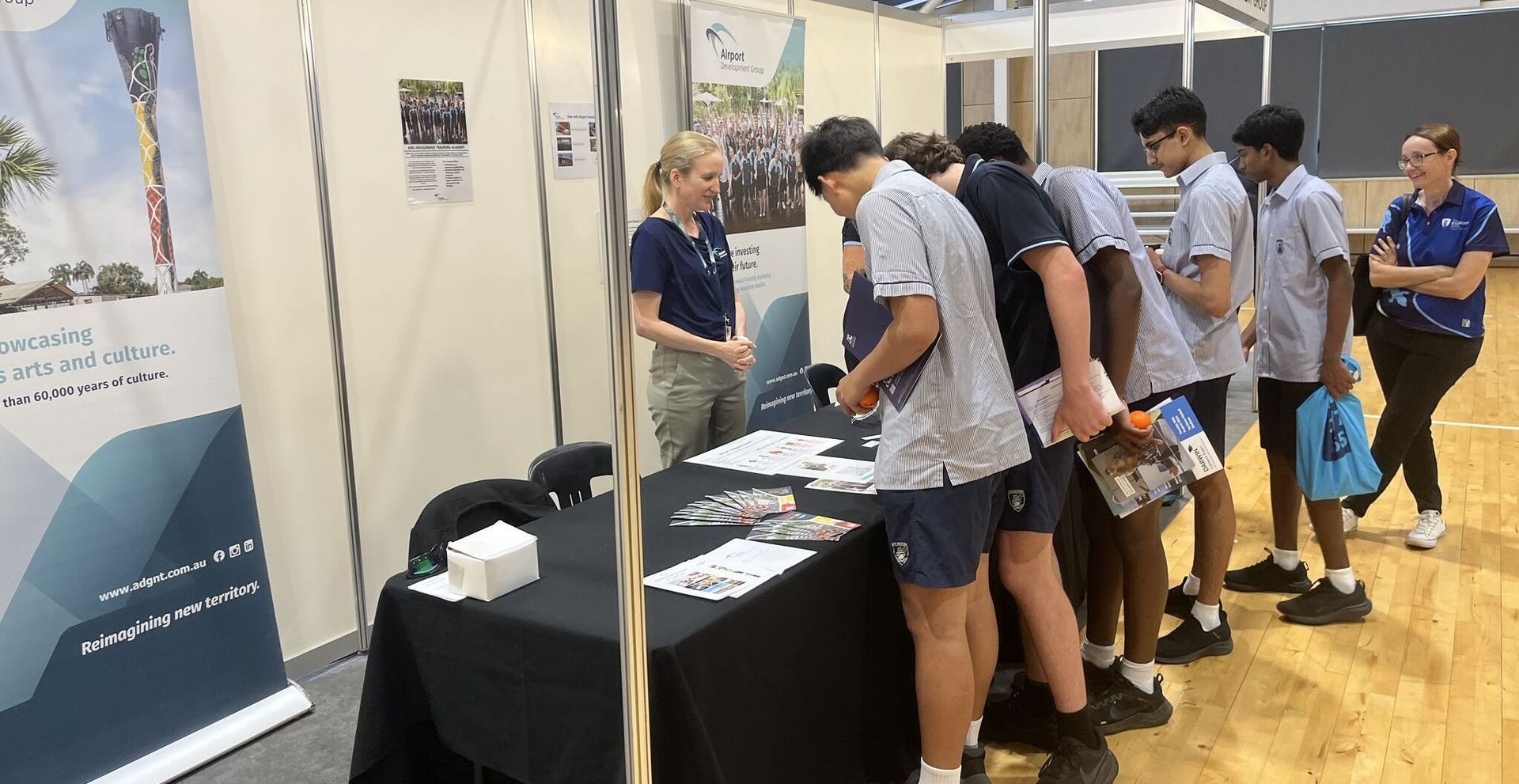 Students at a booth, looking at pamphlets, a woman talking. Gym setting.