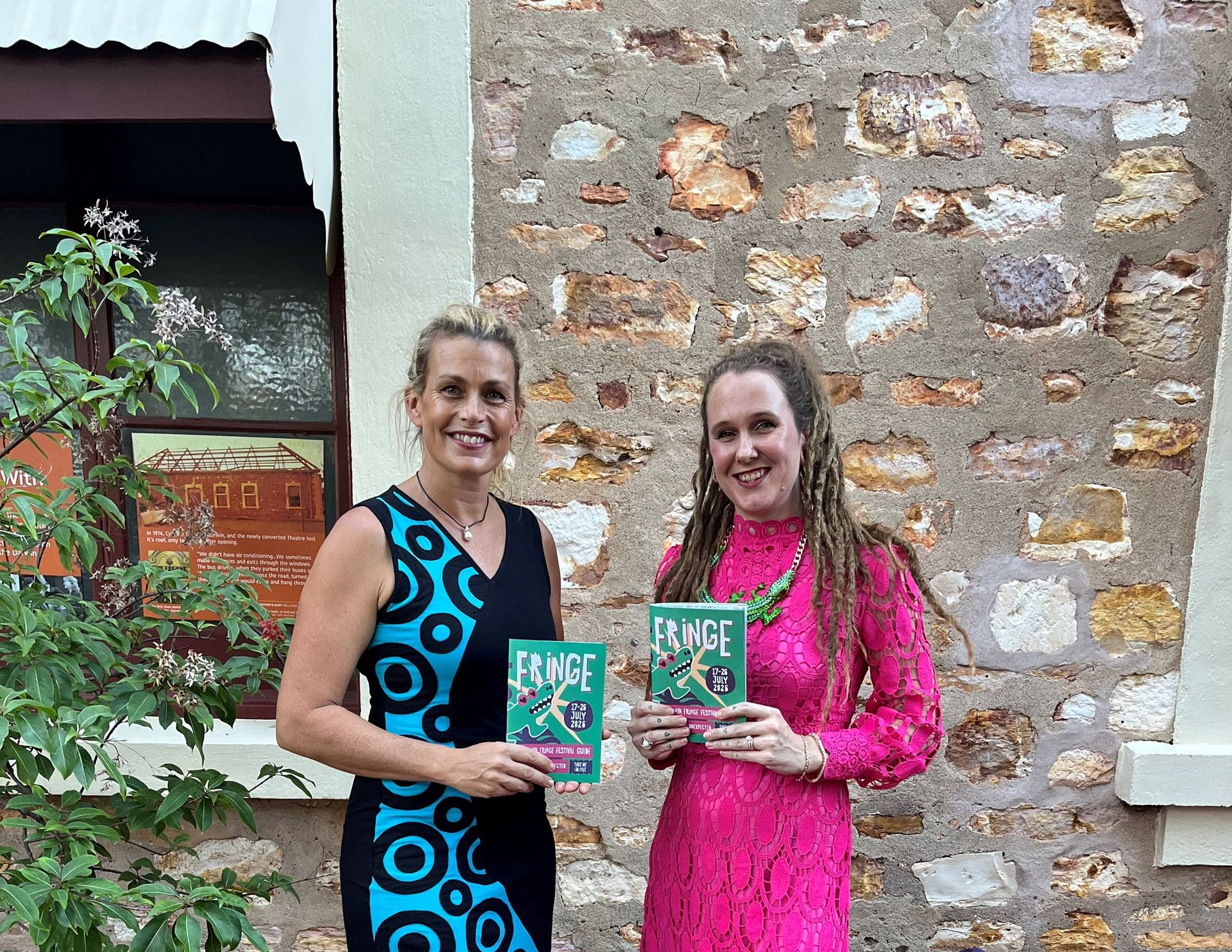 Two women holding a book, standing on a brick patio beside a stone wall.