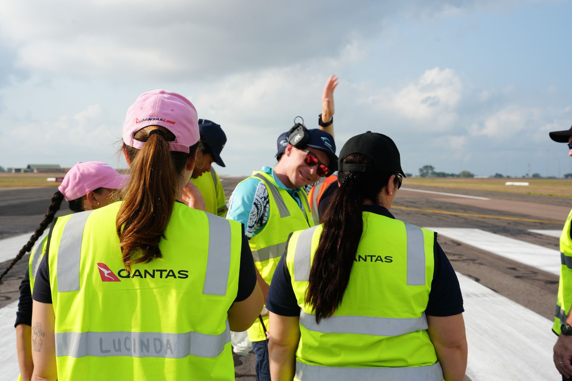 People in neon safety vests and helmets stand on an airport runway, one raising a hand.
