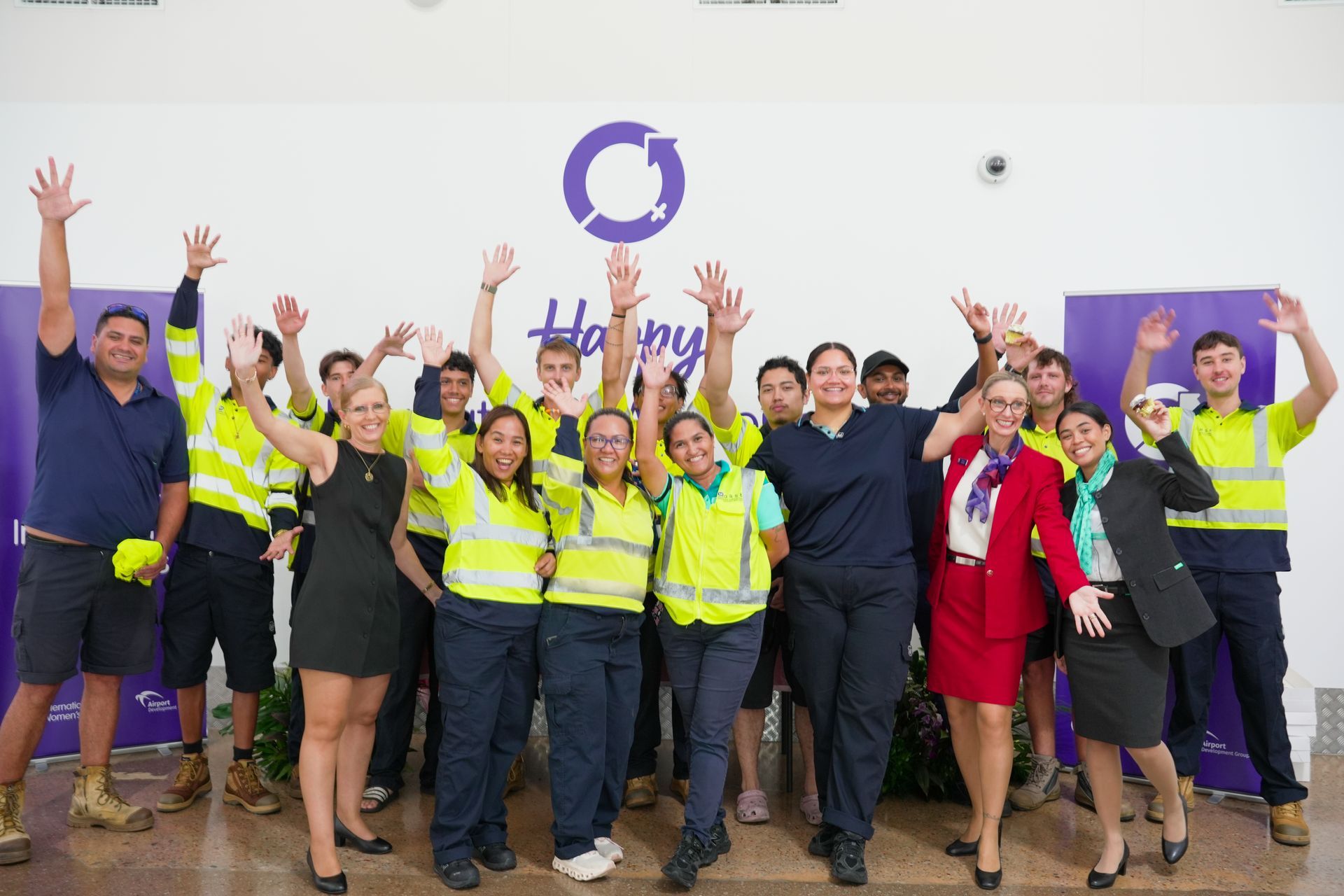 Group of people in various work attire raising their arms, smiling in front of a purple logo and backdrop.