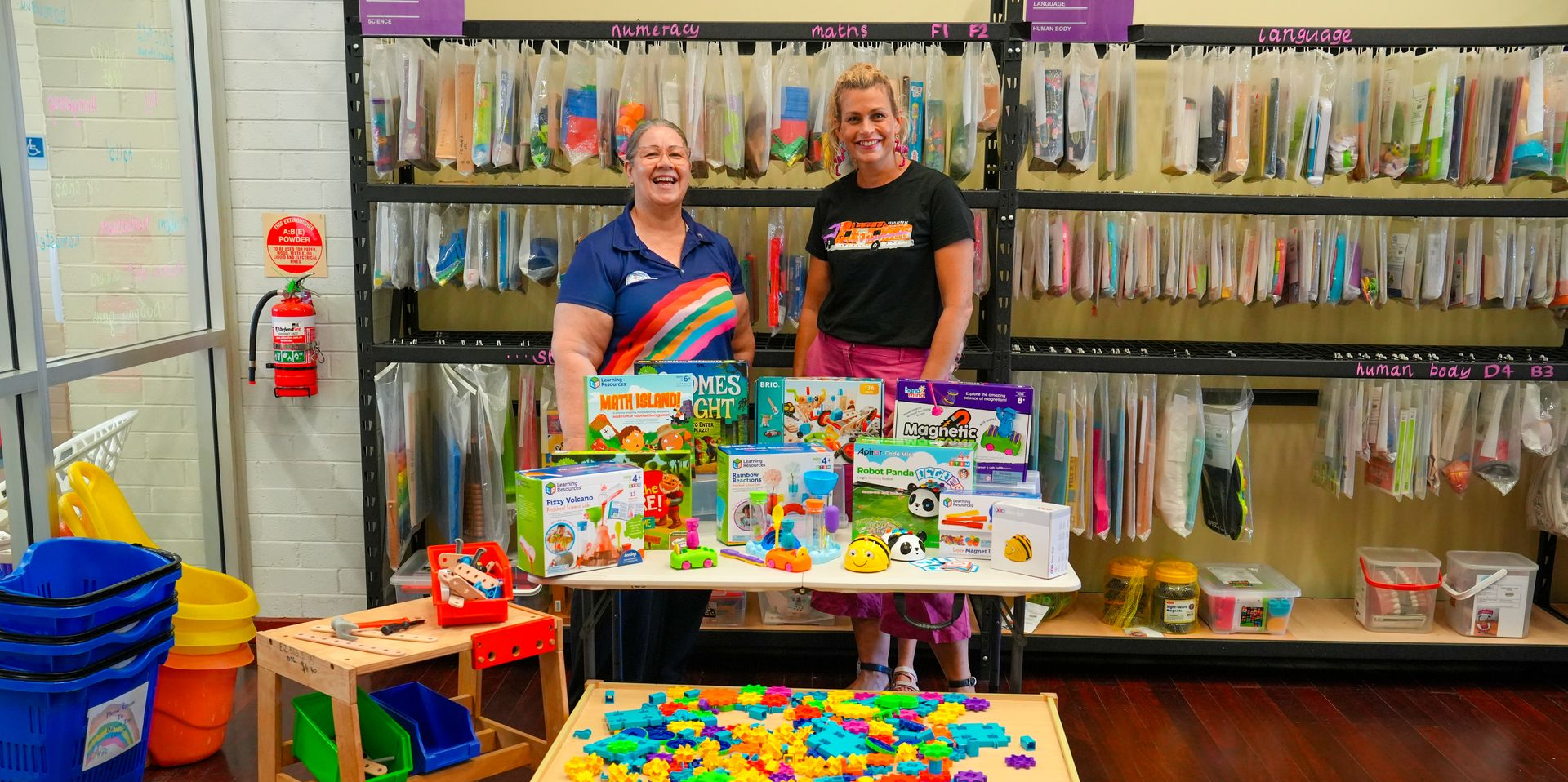 Two people behind a colorful toy display in a store aisle, with shelves of hanging items behind them