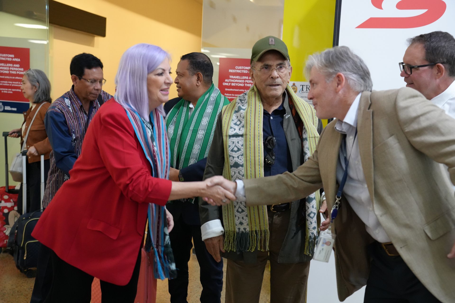 Two people shaking hands at a public event, with others watching in the background.