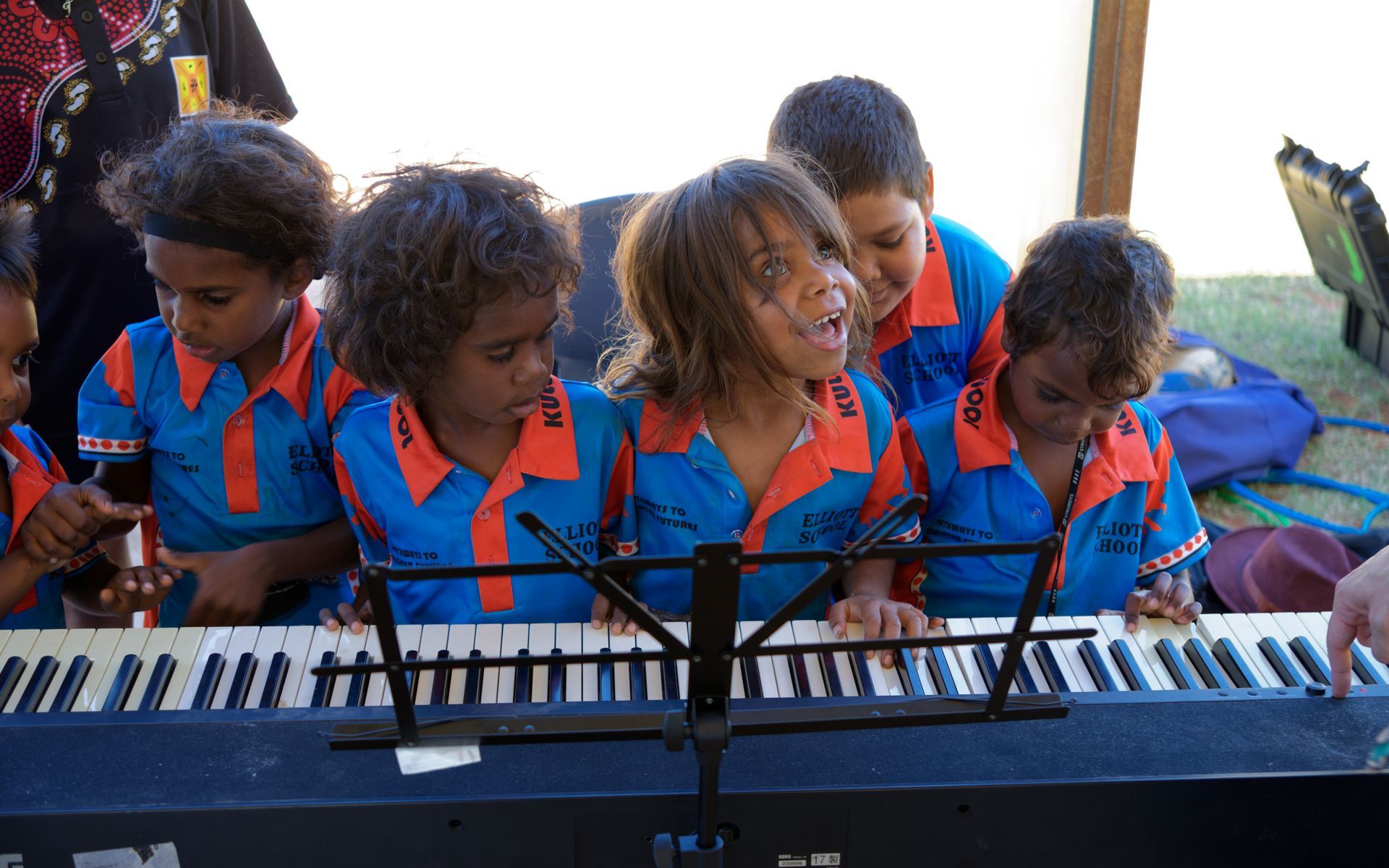Children in blue shirts play a keyboard together outdoors, smiling and singing into microphones.