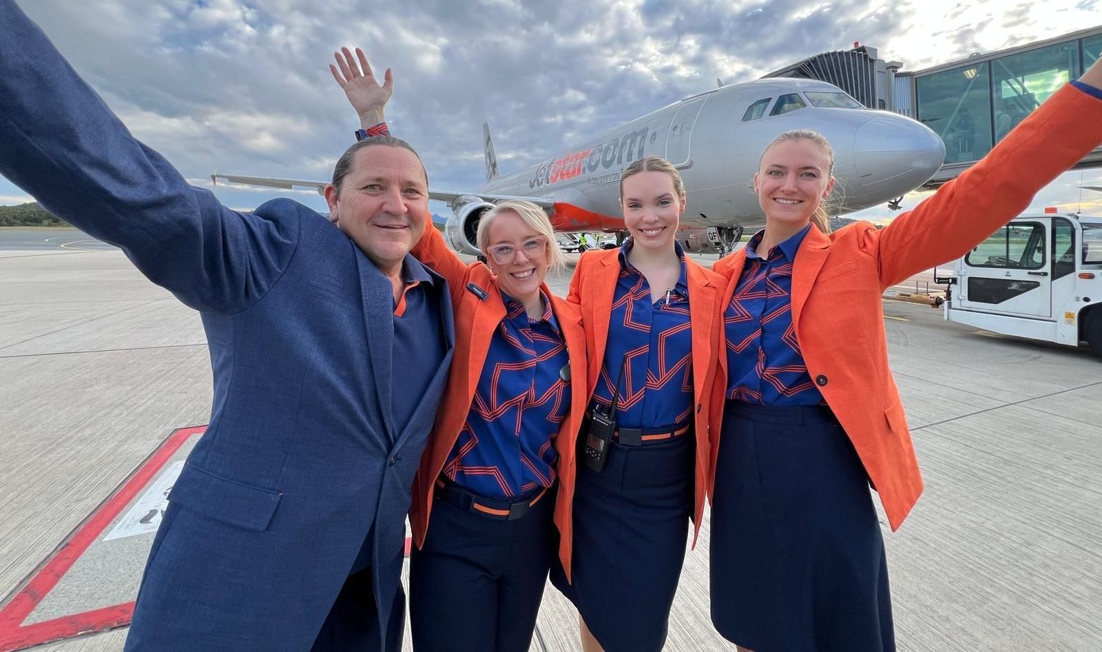 Four airline staff smiling, waving in front of an airplane. They wear orange blazers and blue uniforms.