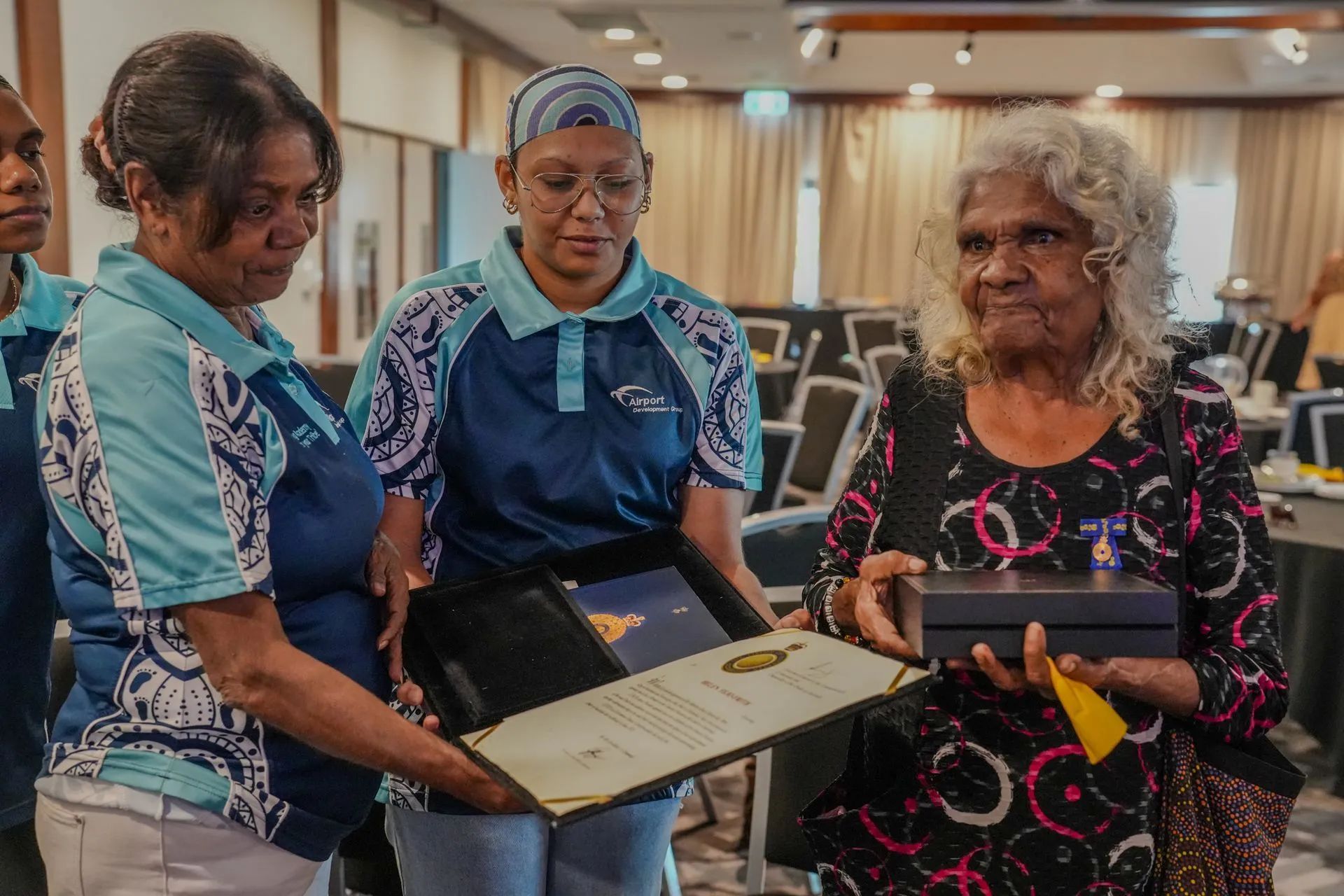 Three women presenting a gift to an elderly woman at an event, indoors.