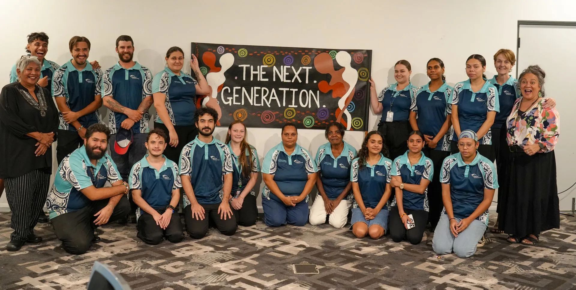 Group of people in matching shirts posing in front of a sign that says "THE NEXT GENERATION."