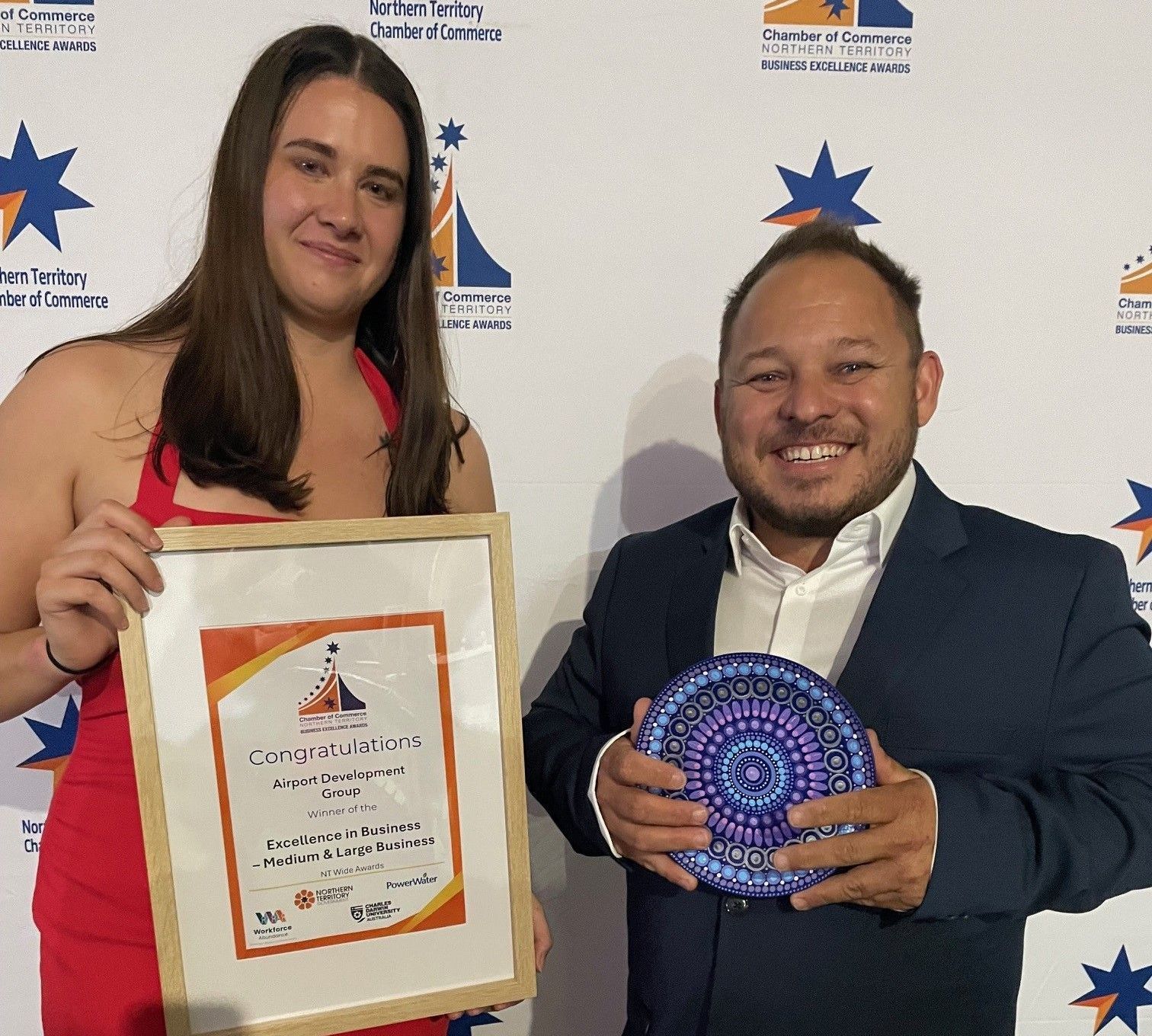 Woman in red dress and man in suit holding award and certificate in front of a logo background.