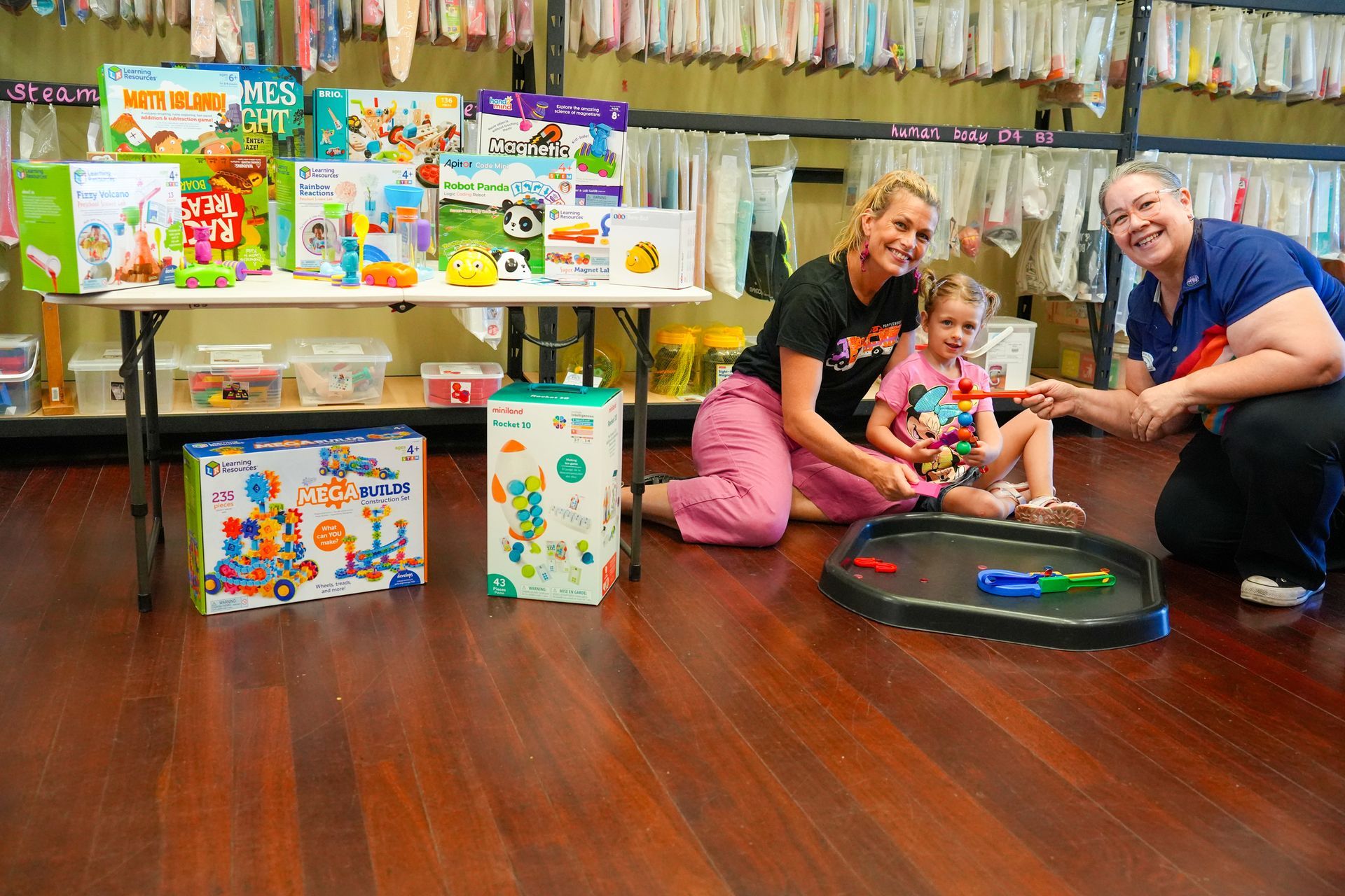 Two people and a child playing with toys on a wooden floor, with toy boxes displayed behind them.