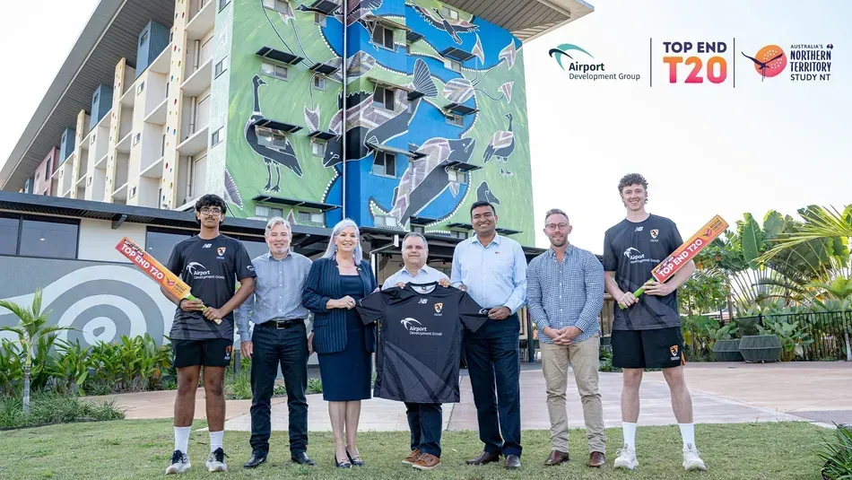 Six people holding cricket bats and a jersey in front of a mural and hotel sign